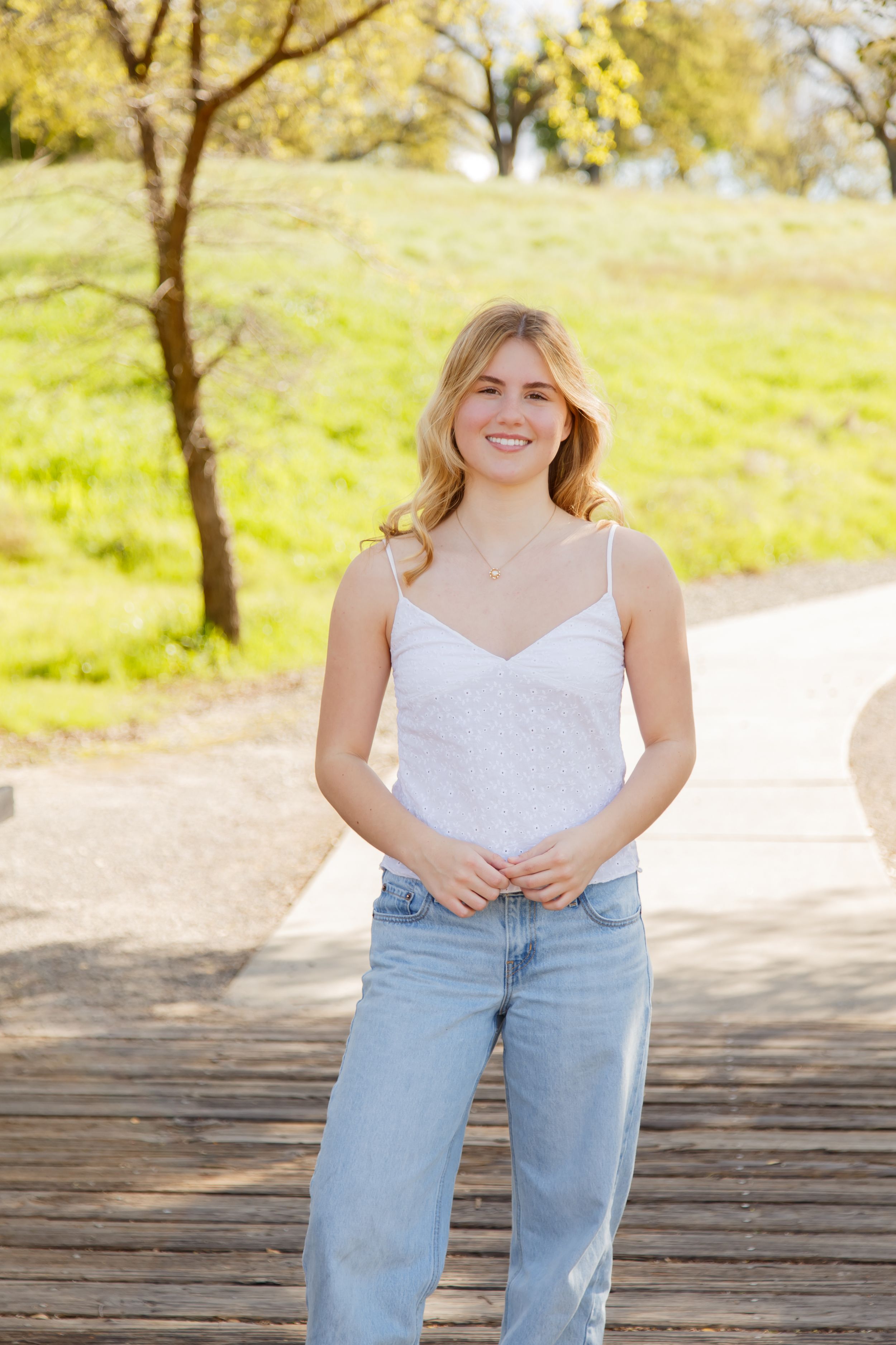 Smiling young woman in white cami top and light blue jeans standing on wooden deck in sunny park.