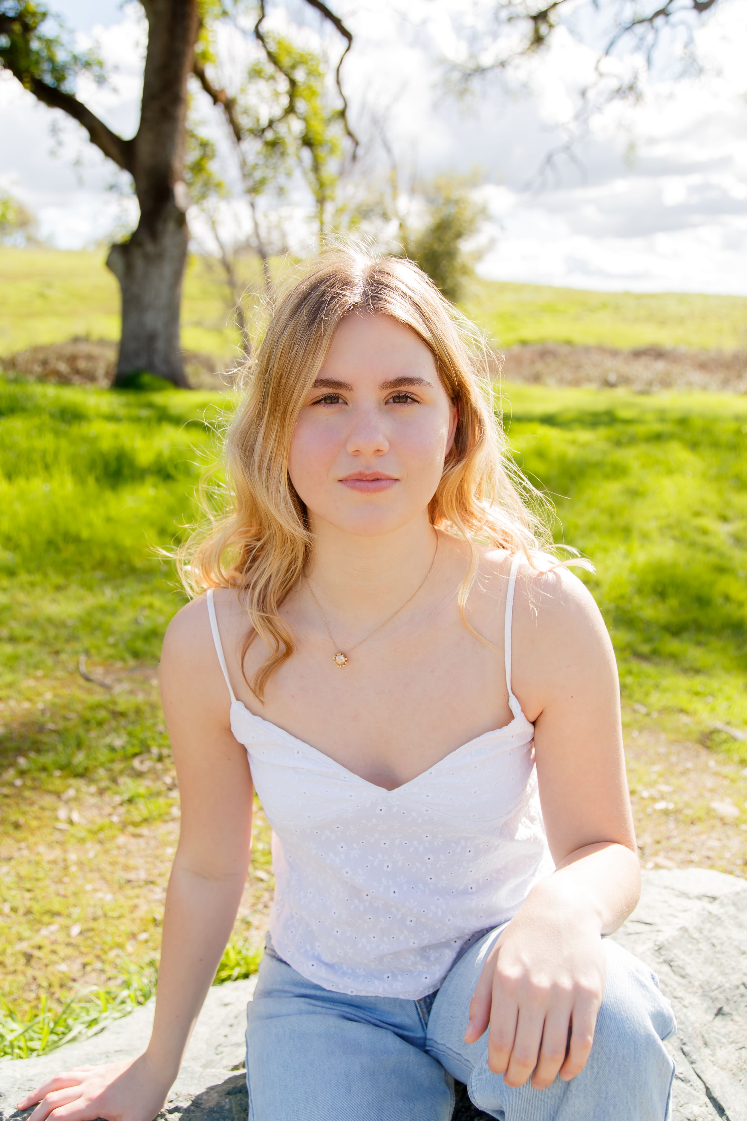 Young blonde woman in white tank top and jeans sitting outdoors on a sunny day with green hills and tree.