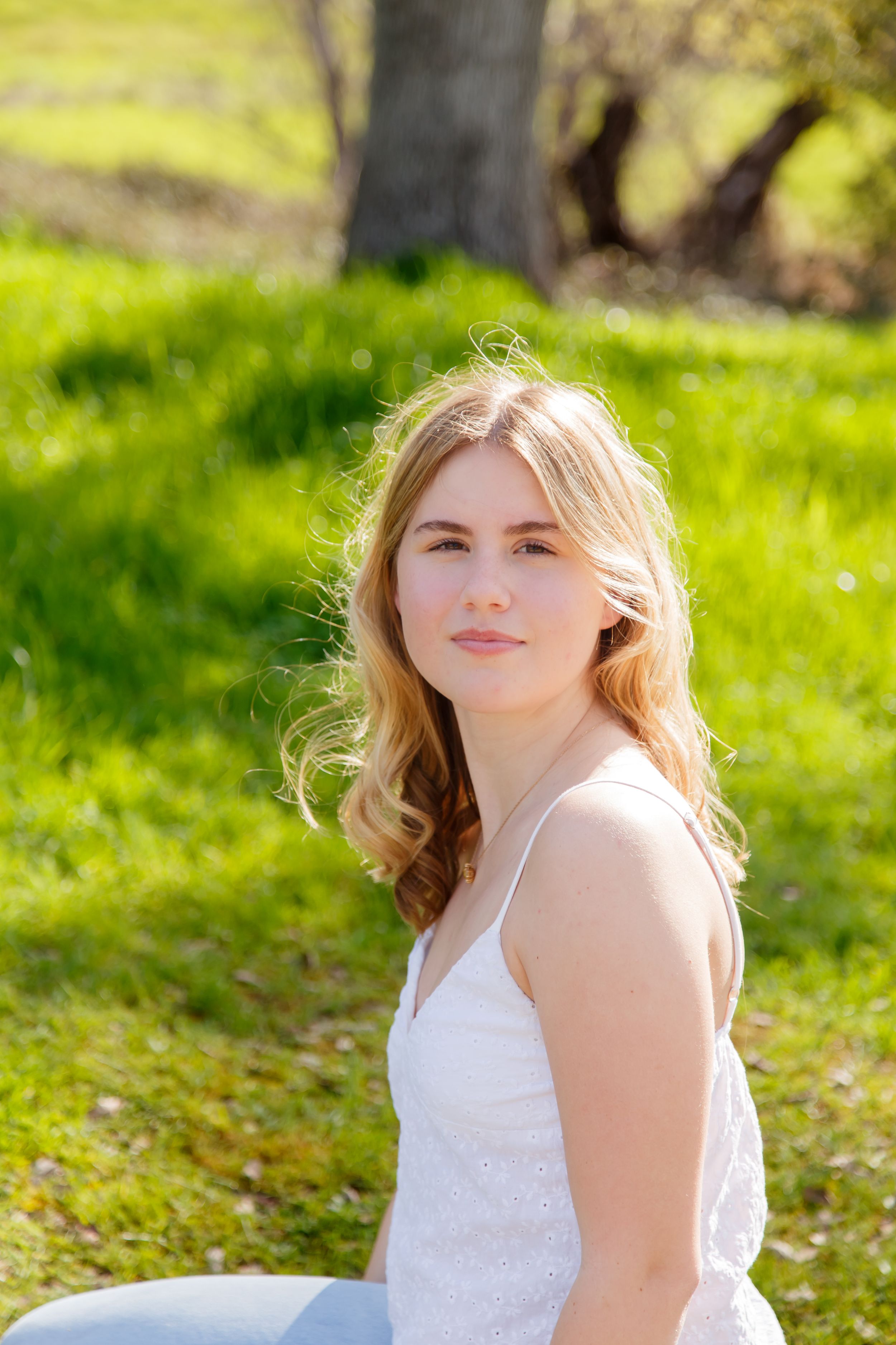 Young blonde woman in white sundress sitting outdoors on green grass in sunlight.