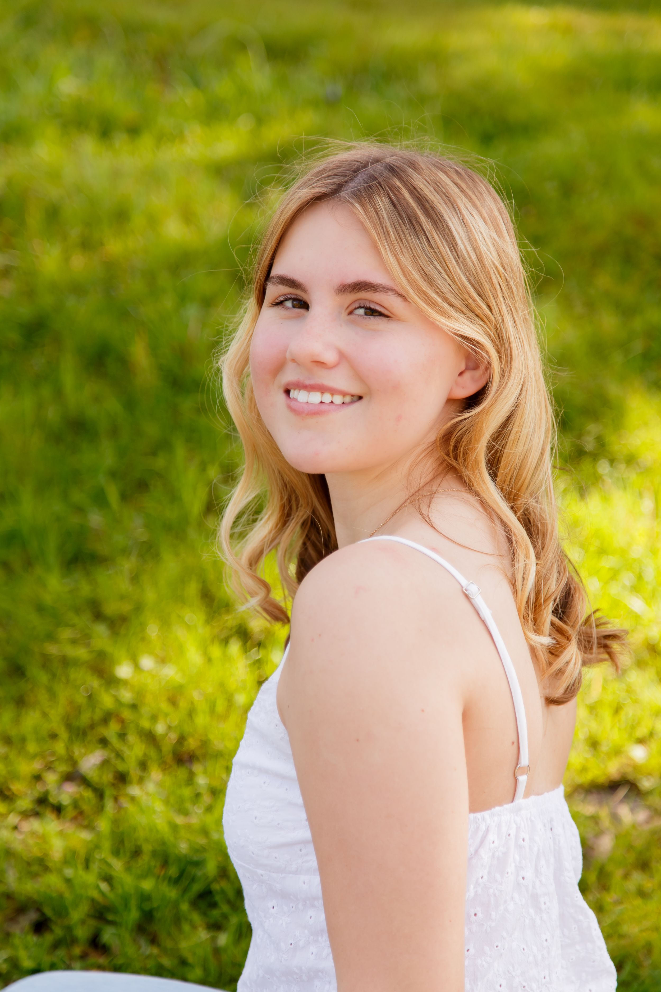 Smiling blonde teen girl in white sundress sitting on green grass in sunny park.