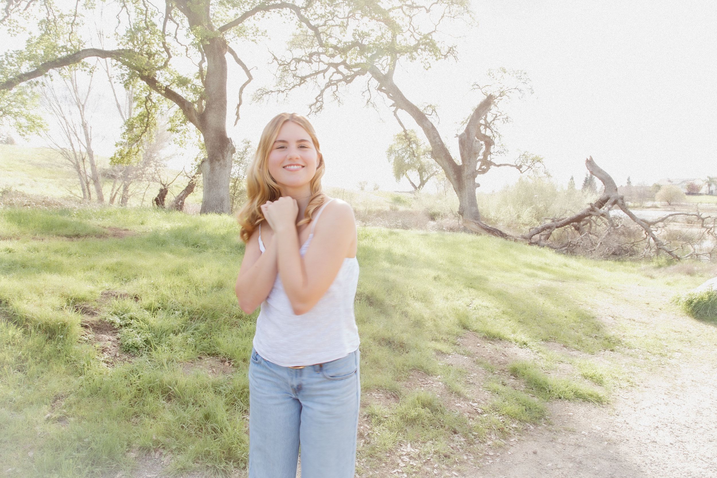 Smiling young woman in white tank top and jeans standing on a dirt path surrounded by green trees outdoors.
