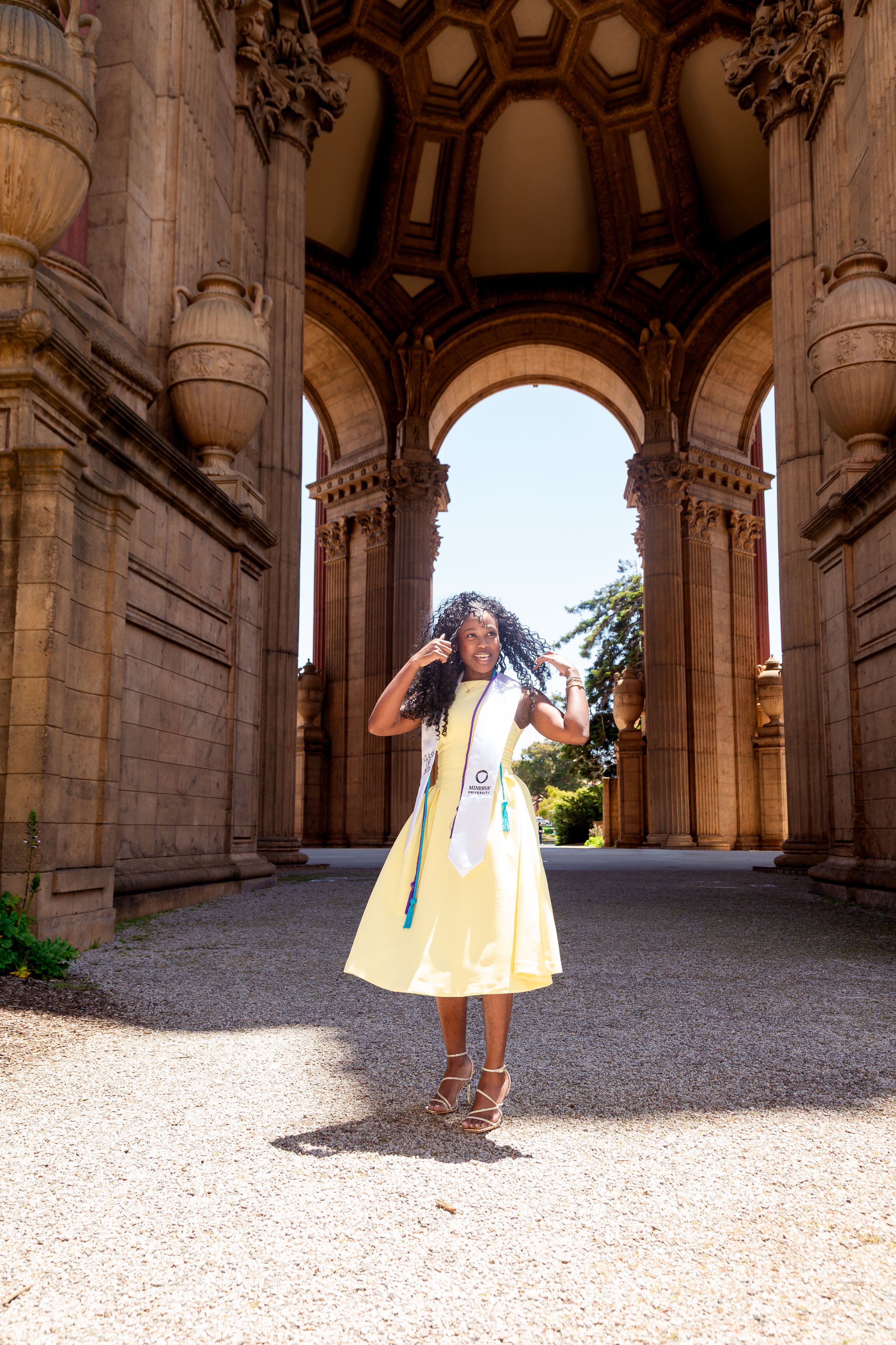 A graduate in a yellow dress and stole poses joyfully at the Palace of Fine Arts in San Francisco.