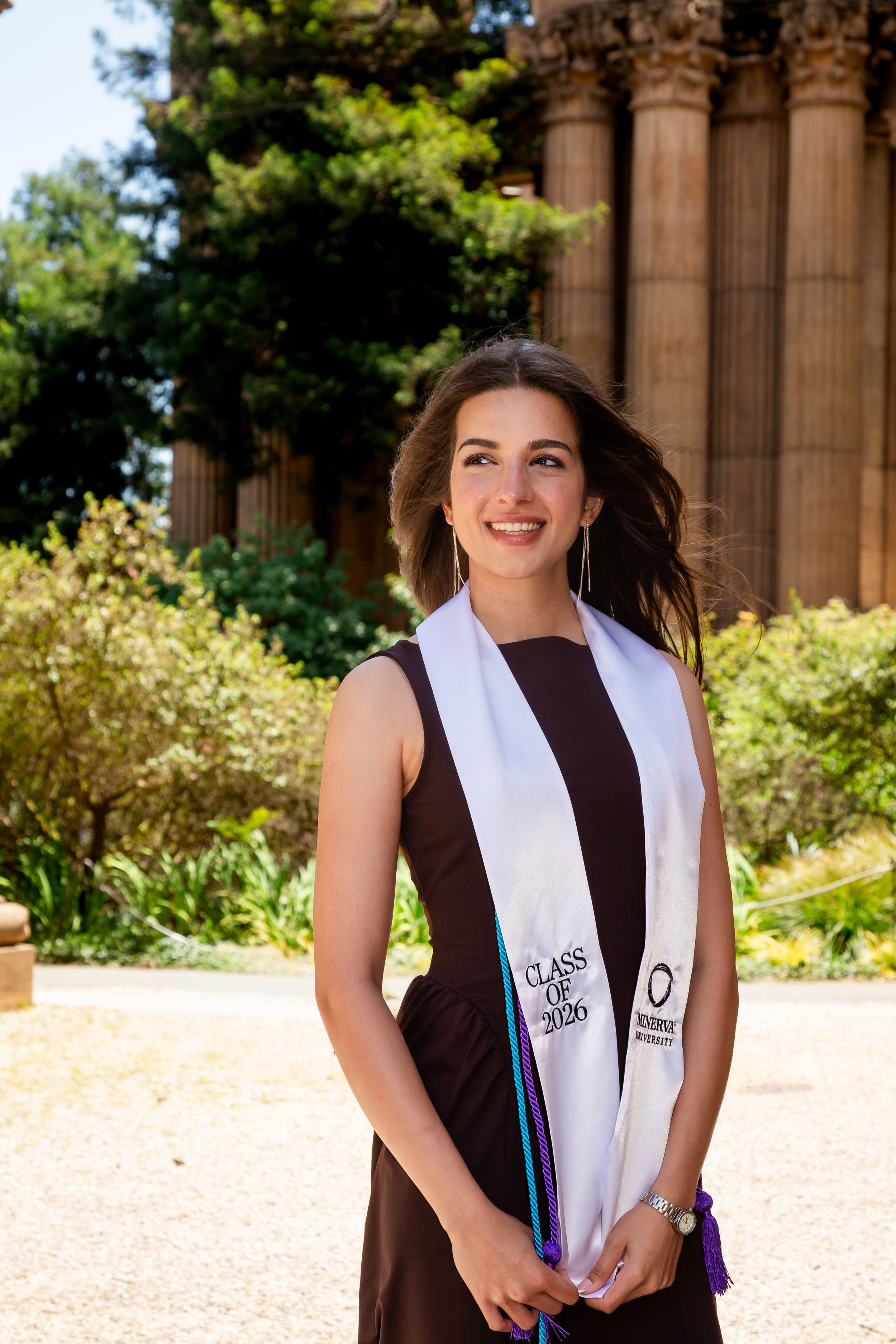 Smiling young woman in brown dress wearing graduation stole outdoors near classical columns.