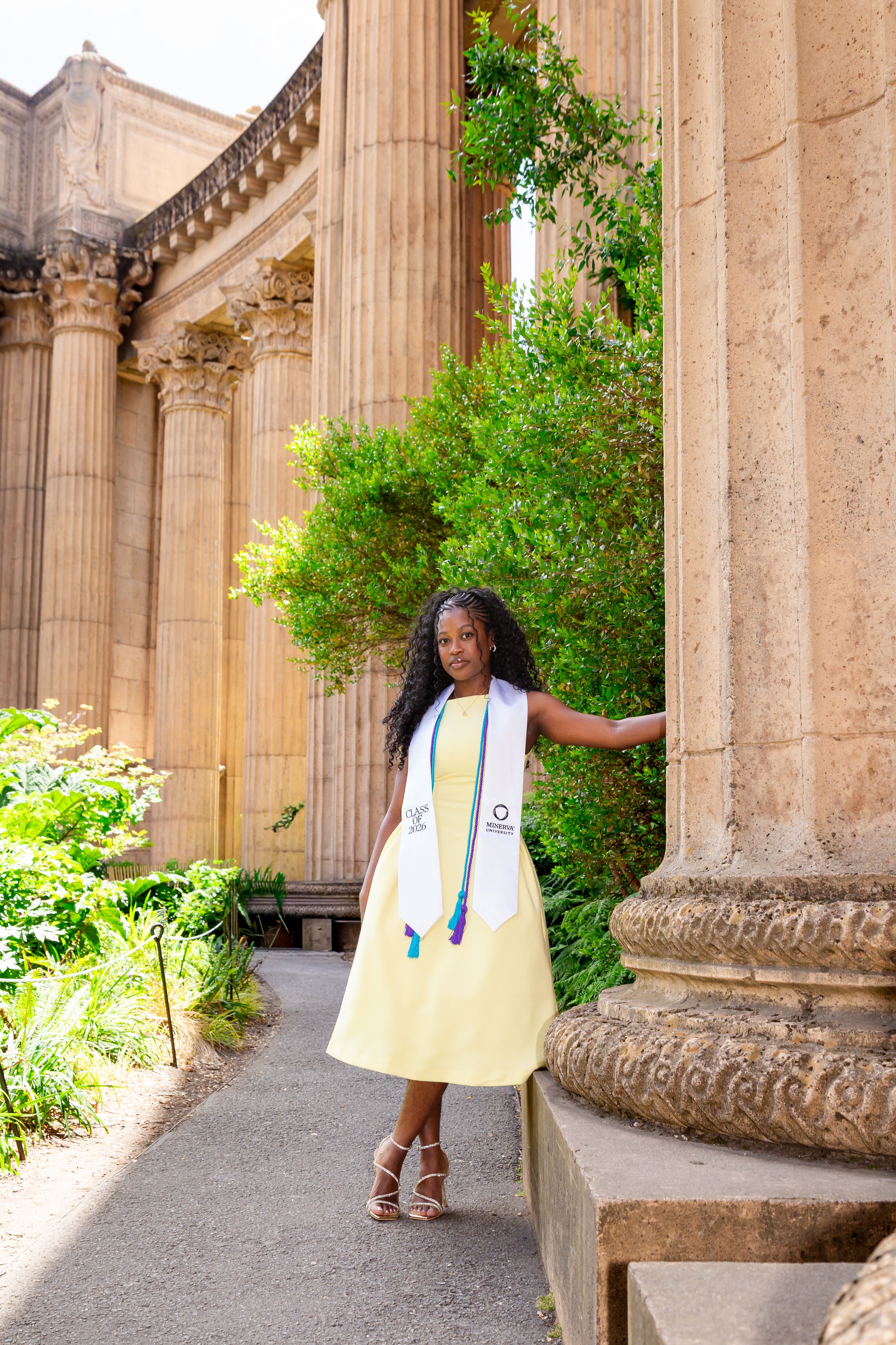 Young woman in yellow graduation dress and stole poses between ornate columns at Palace of Fine Arts.