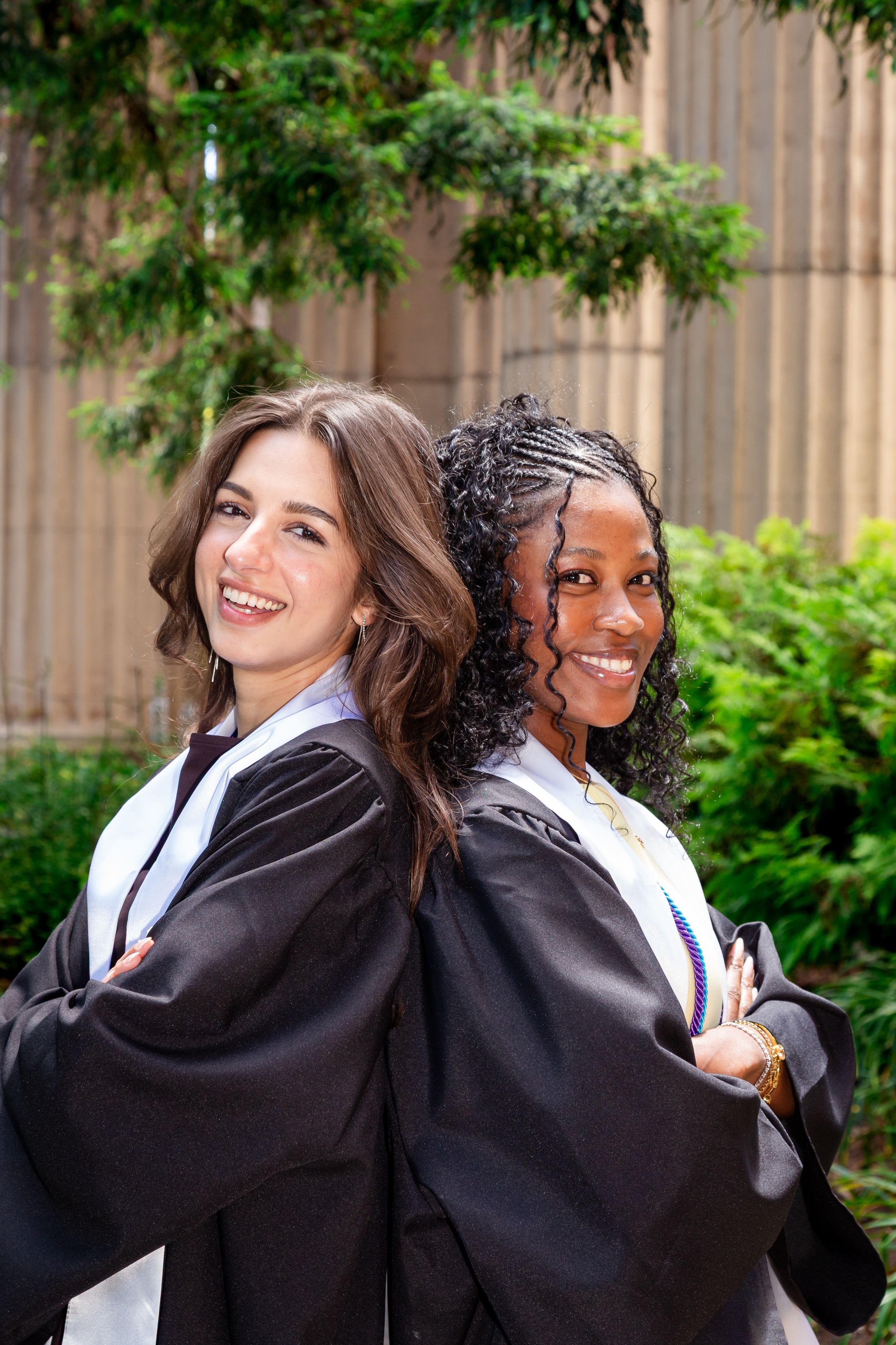 Two smiling graduates in black gowns and white stoles pose back-to-back outdoors among green foliage.