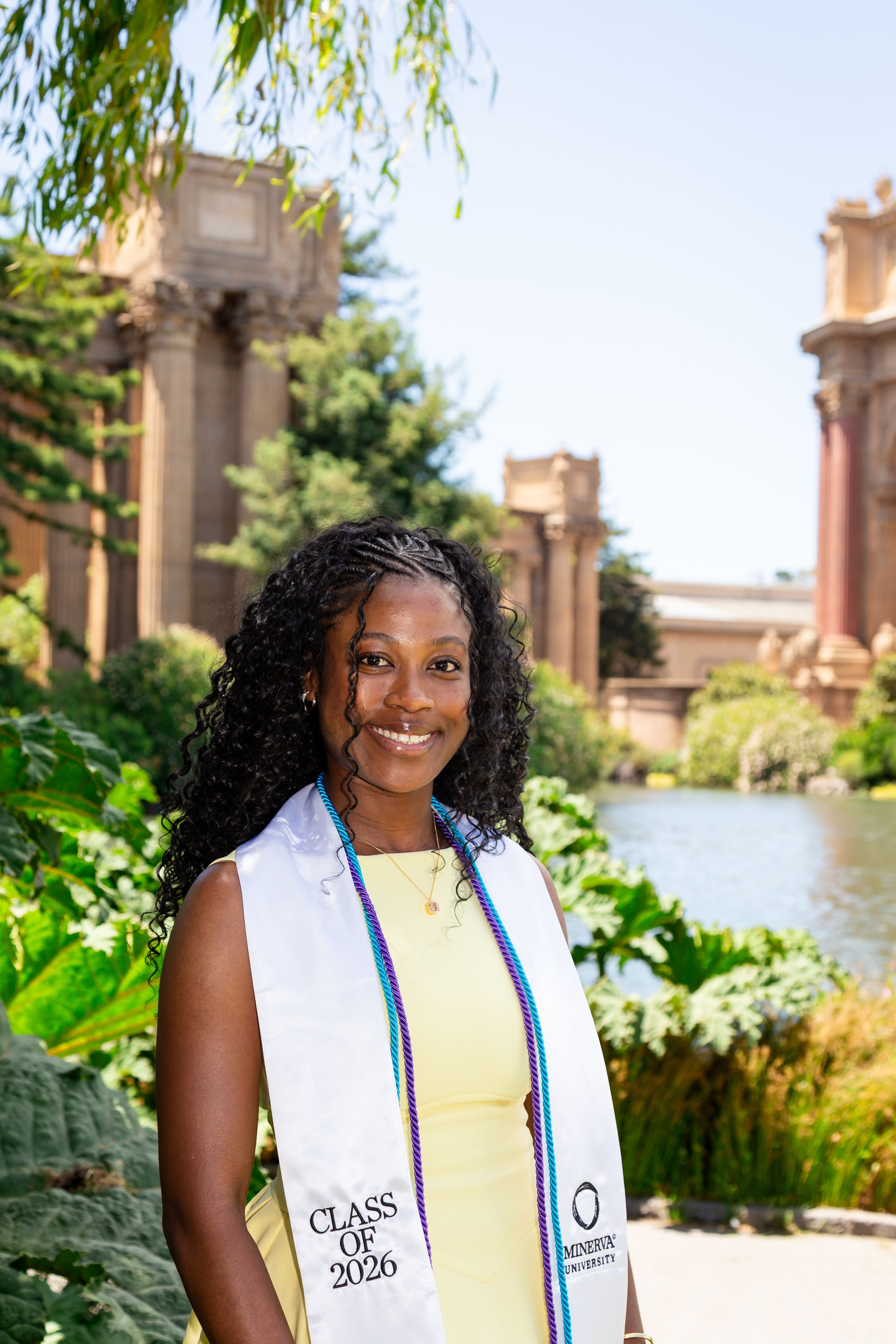 A smiling graduate wearing a Class of 2026 stole poses outdoors near a pond with classical architecture behind her.