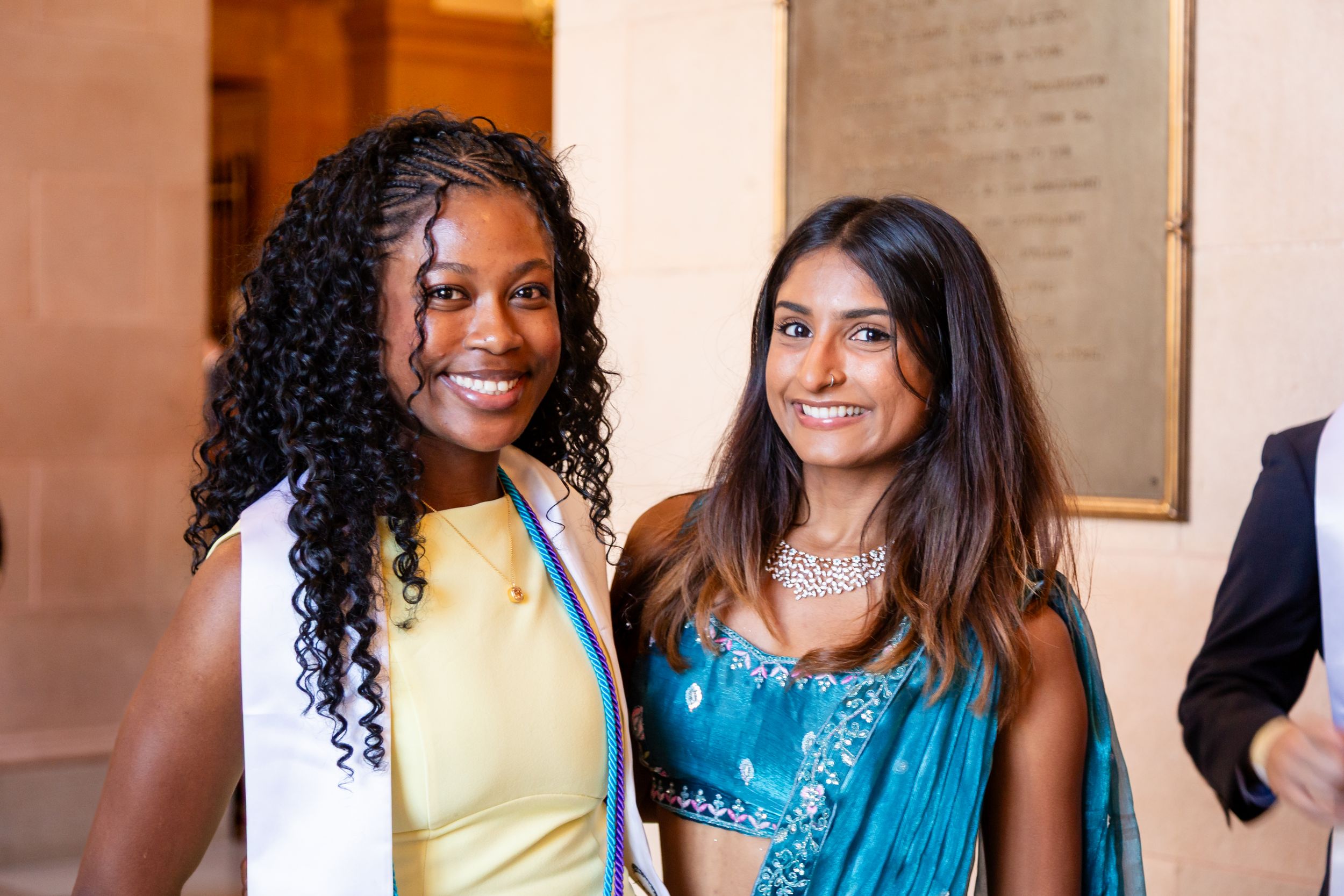Two women smiling at a formal event, one in a yellow dress and one in a blue embroidered sari with jewelry.