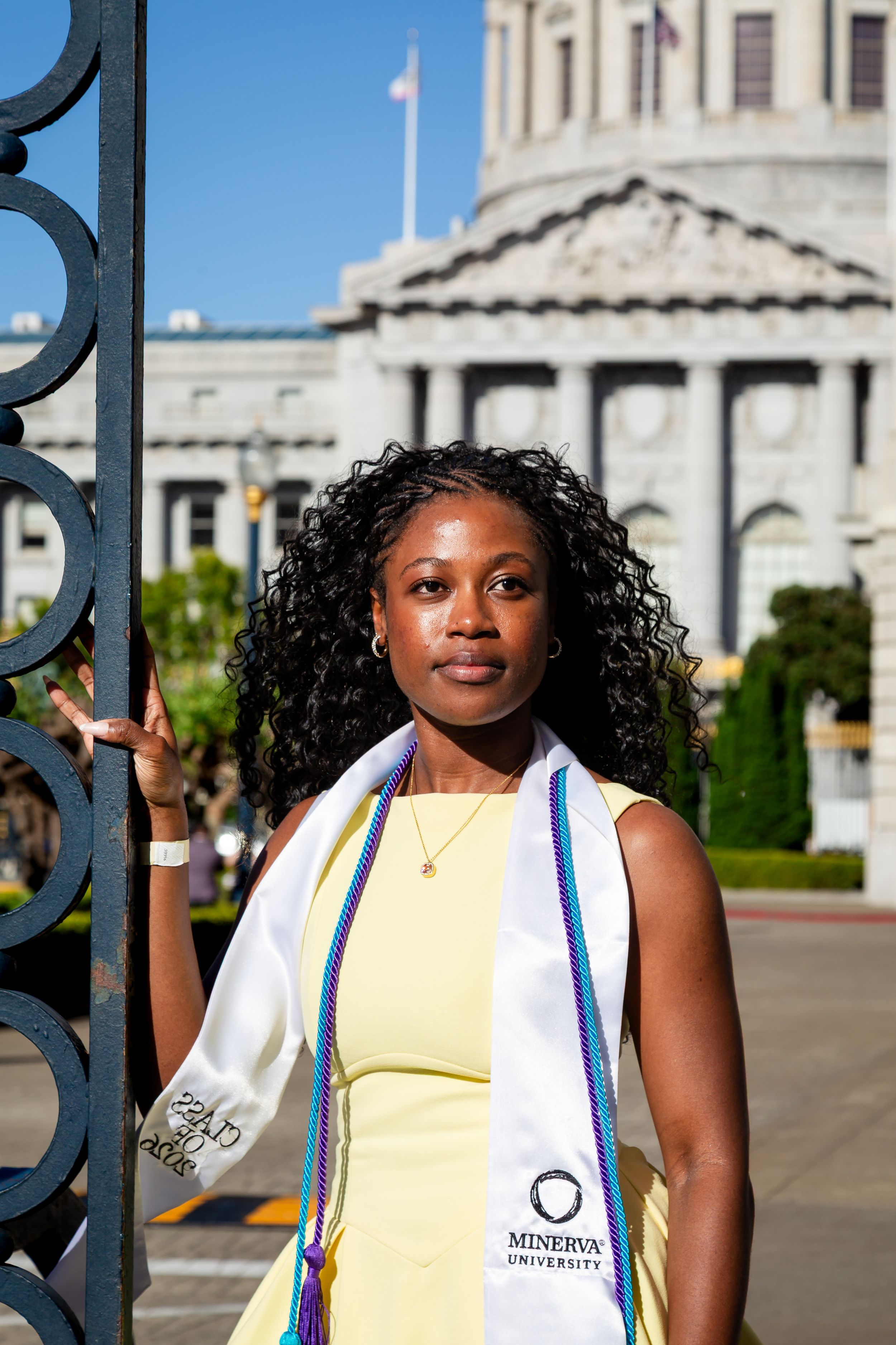 A Minerva University graduate in a yellow dress and white stole poses by iron gates with a classical building behind her.