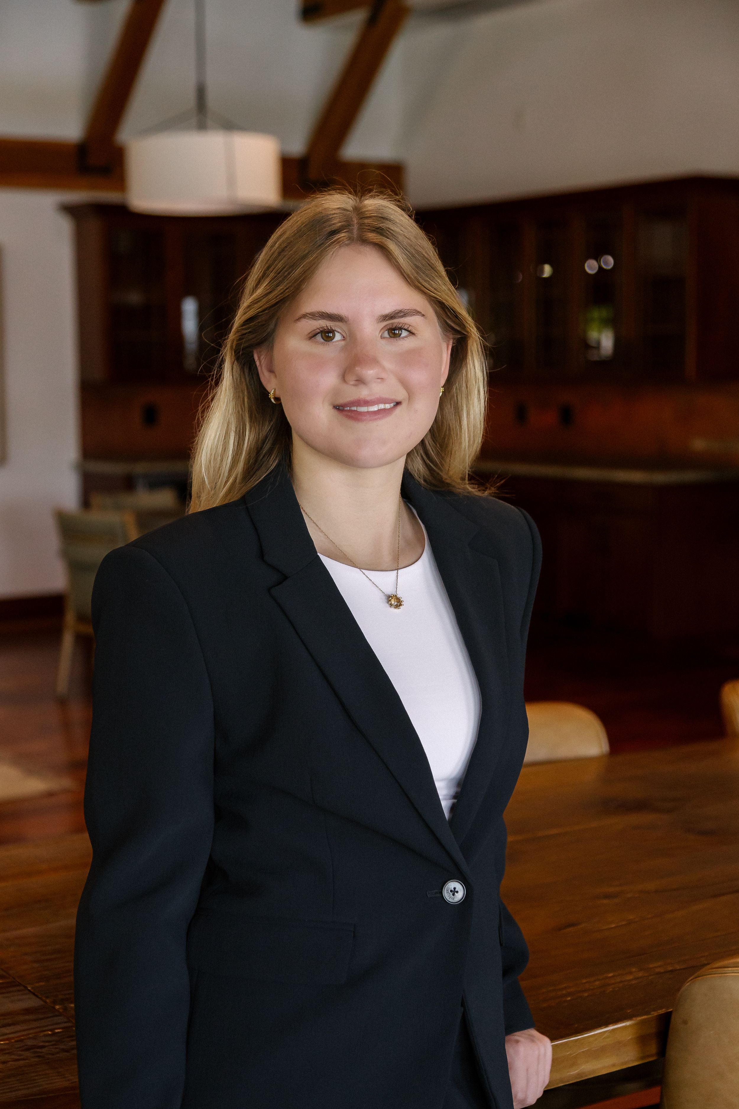 Young professional woman in black blazer smiling in elegant wood-paneled dining room setting.