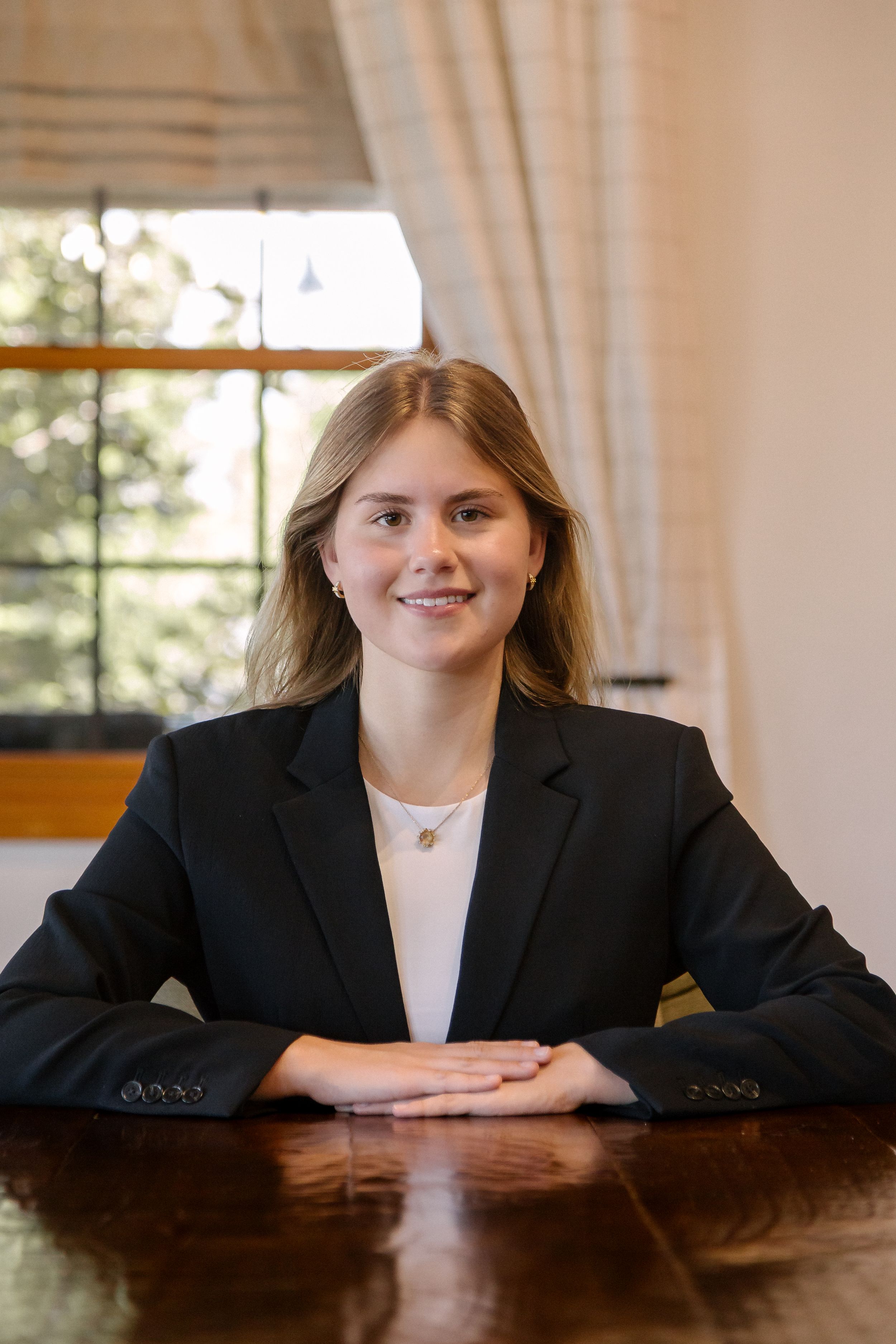 Young professional woman in black blazer smiling at wooden table in bright office setting.