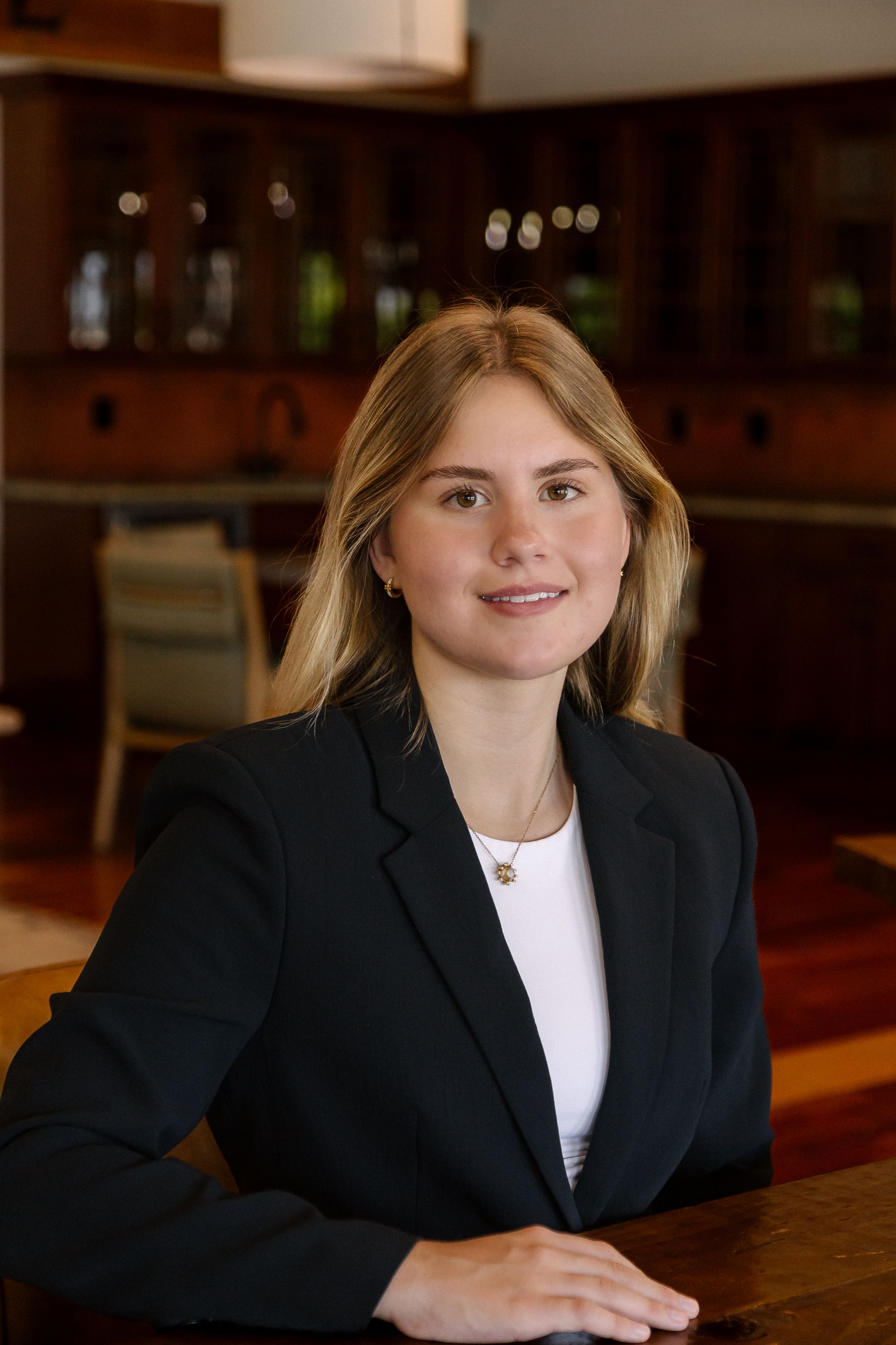 Blonde professional woman in black blazer posing at wooden table in upscale interior.