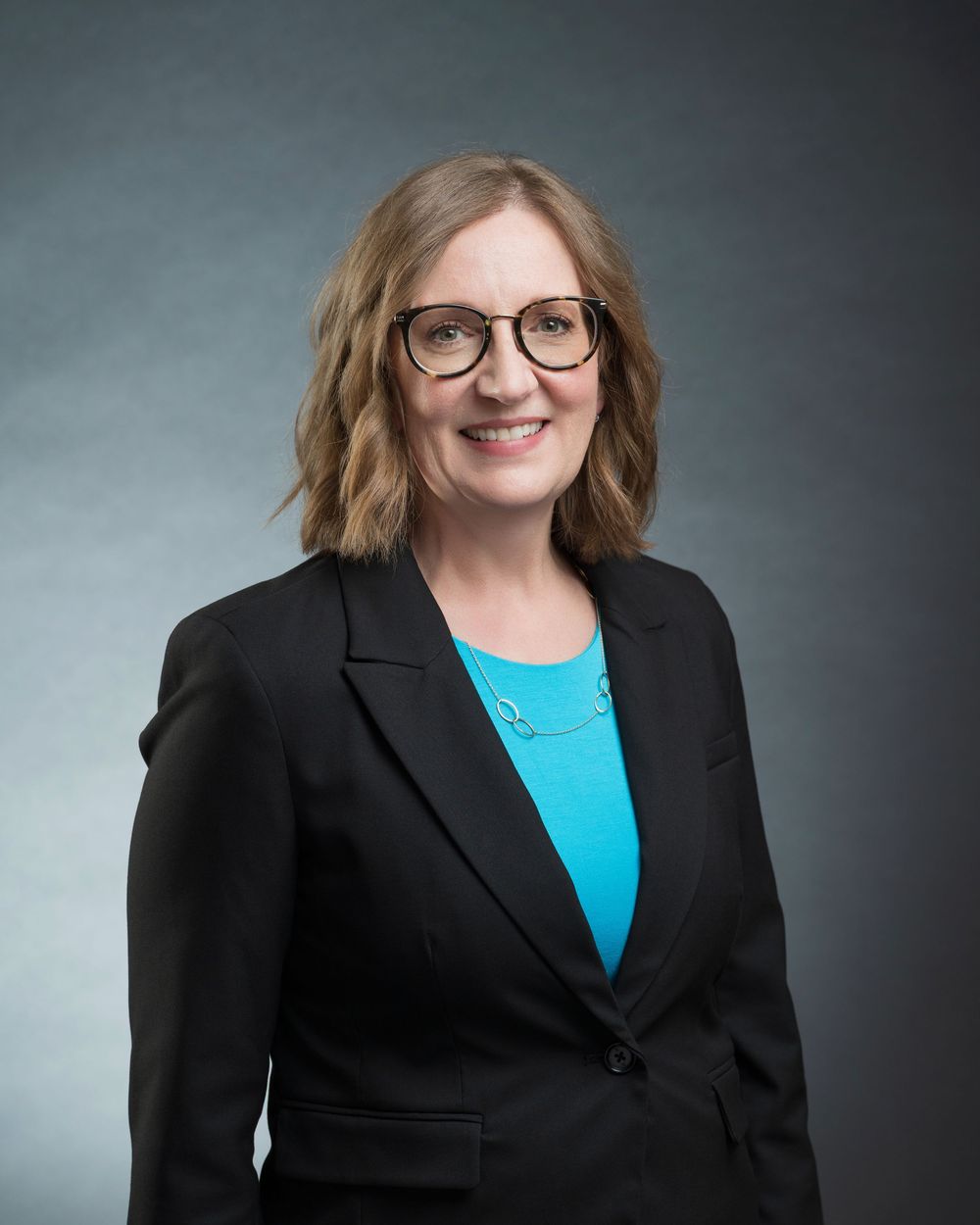 Professional woman in black blazer and teal top with glasses, smiling confidently against gray background.