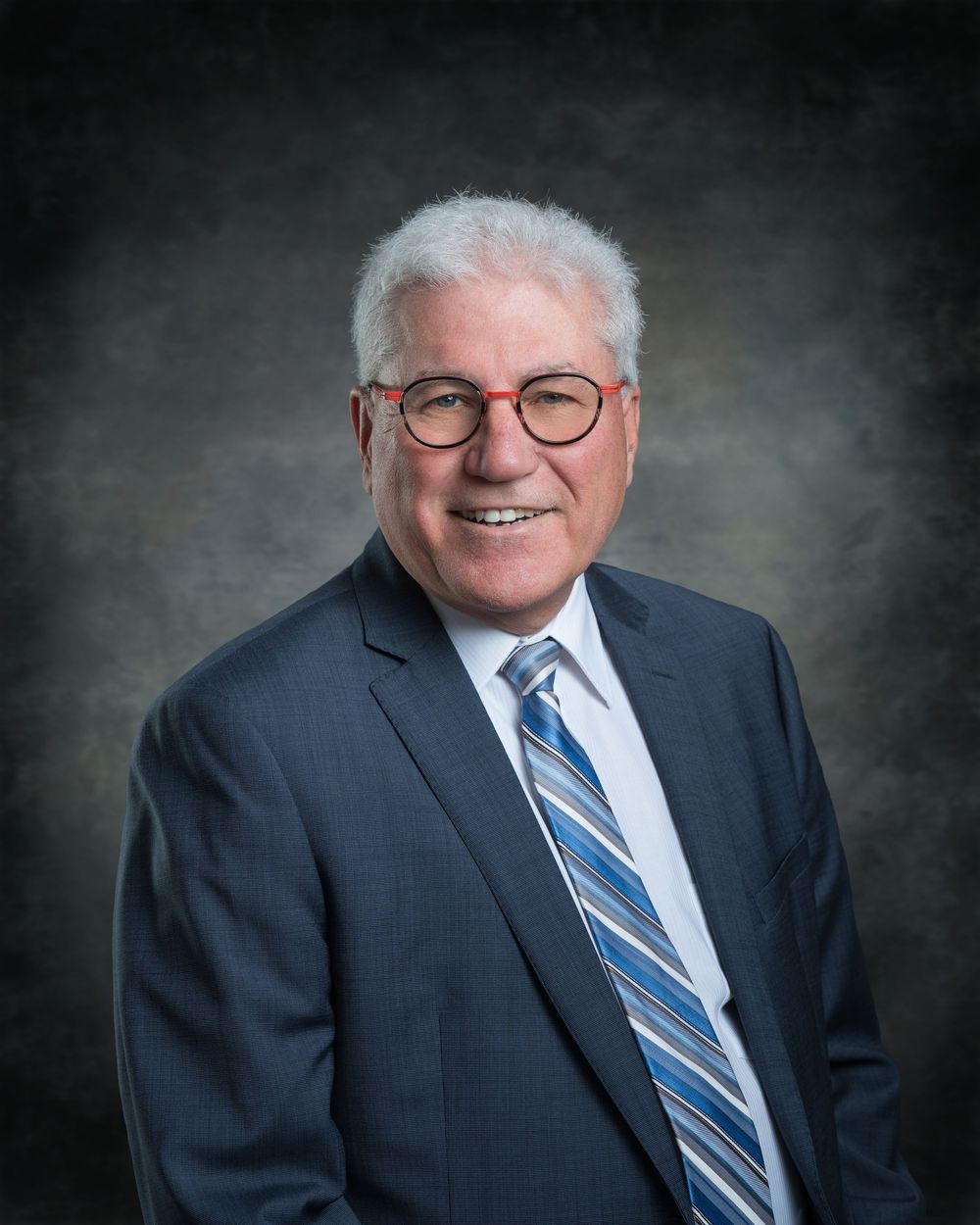 Professional headshot of a smiling senior businessman with white hair and glasses wearing a blue suit and striped tie.
