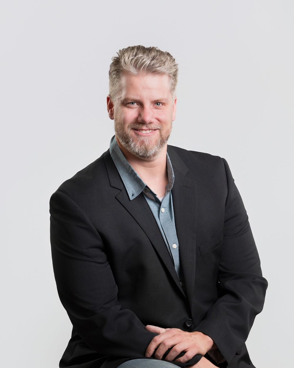 Professional man with blonde hair and beard wearing a black blazer, smiling confidently against a gray background.