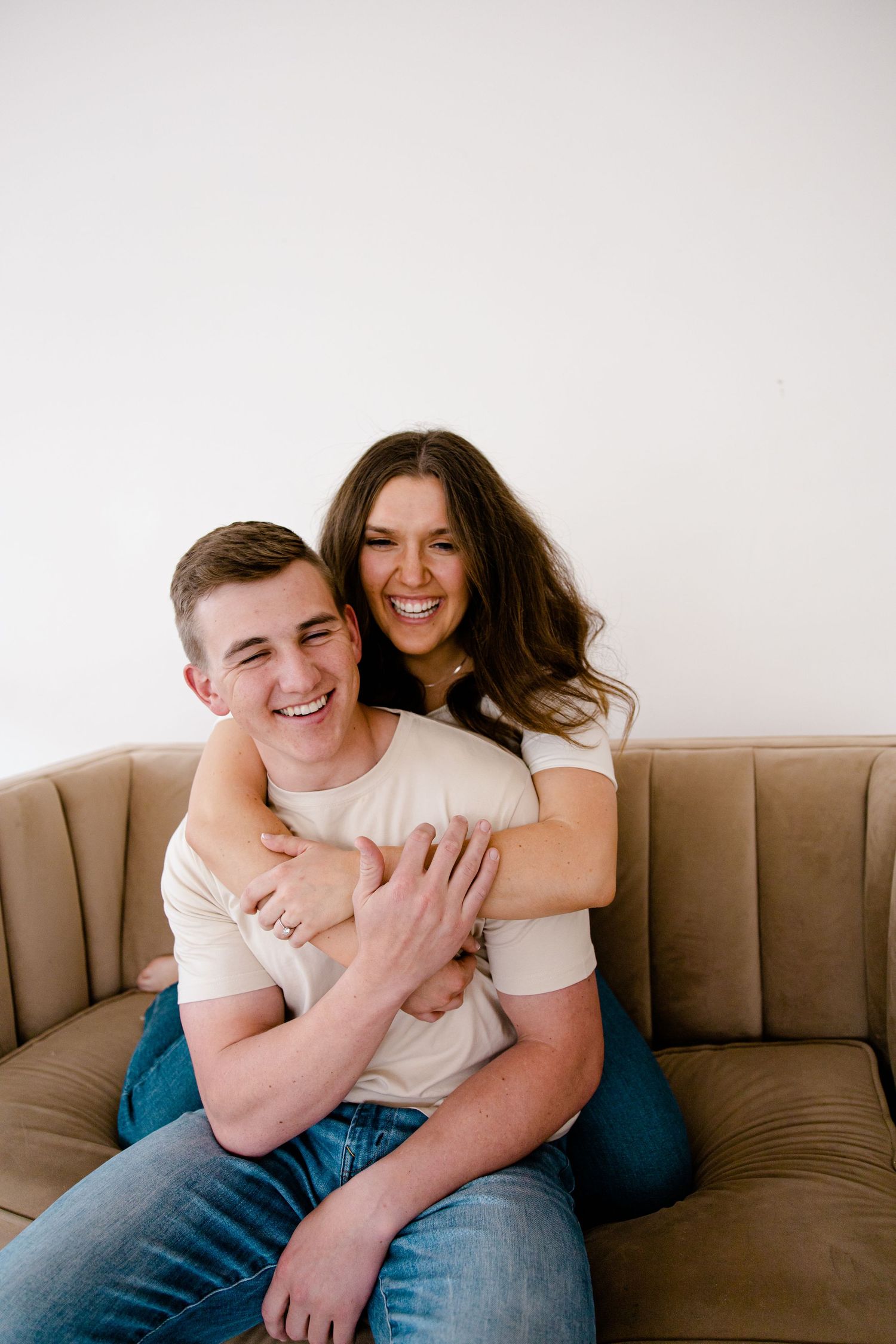 Sioux City, Iowa photographer. Smiling couple hugging on a beige couch, woman embracing man from behind joyfully.