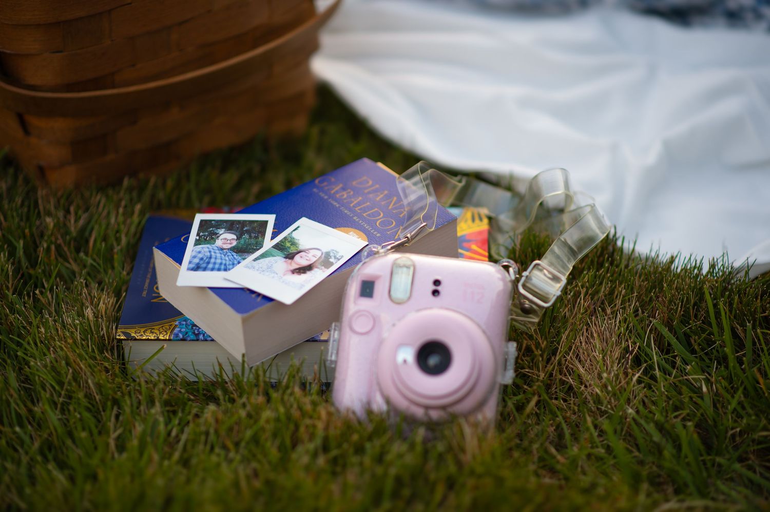 Pink Instax Mini camera beside books and picnic items on green grass outdoors.