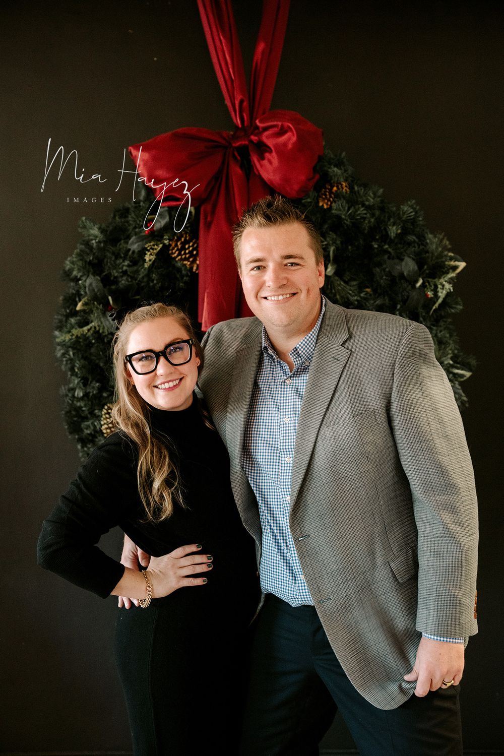 Couple posing for professional photos in front of Christmas wreath and dark backdrop.