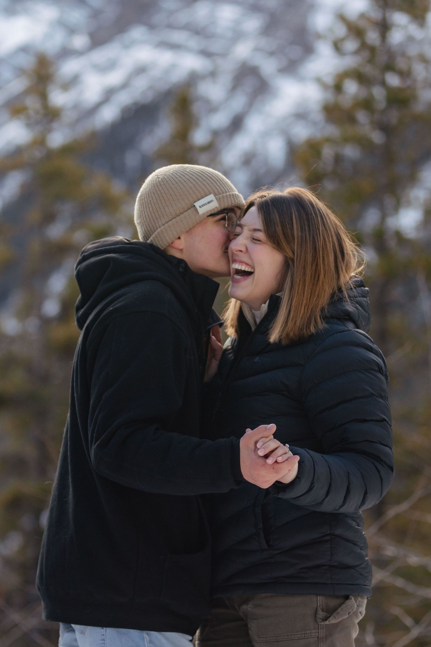 A couple dance in a wooded, winter, mountain landscape. The man is whispering in the woman's ear. The woman is laughing.