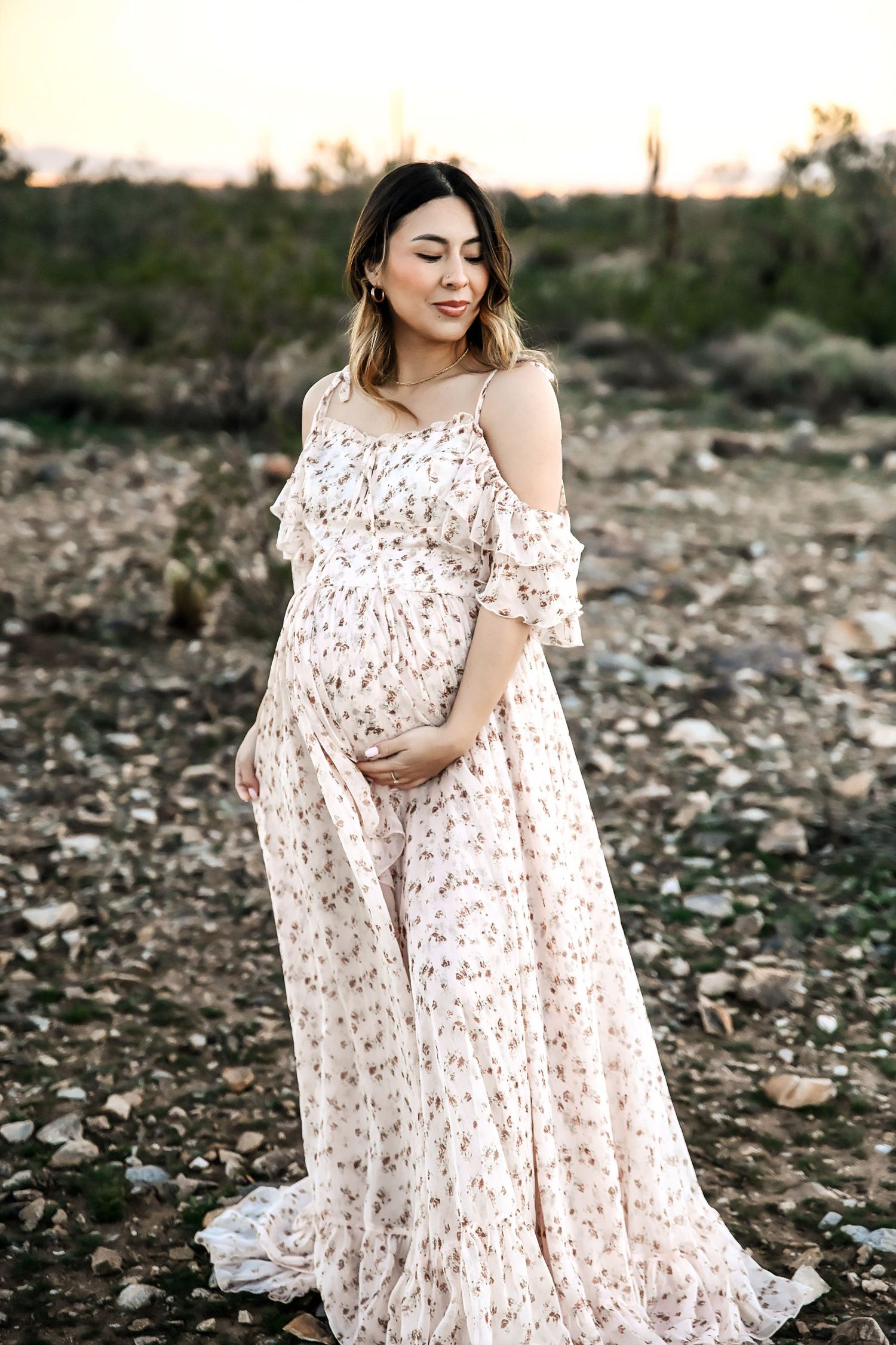 Pregnant woman in floral maxi dress cradling baby bump outdoors at sunset in a rocky desert landscape.