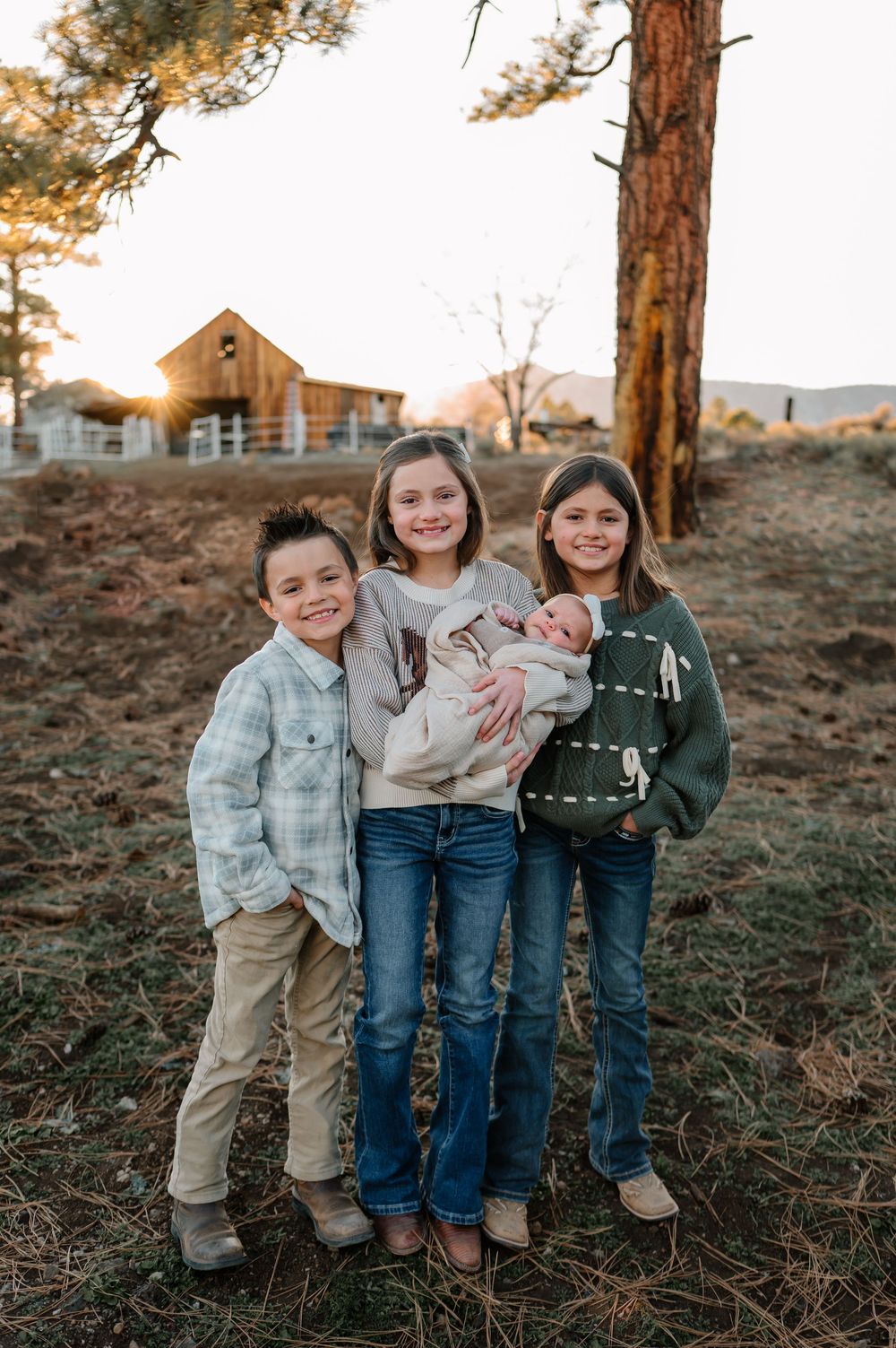 Three children stand together among tall pine trees at sunset in casual fall clothing.