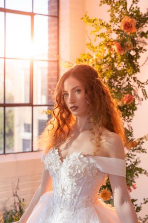 A bride in an ornate white off-shoulder wedding gown with floral lace details poses in warm sunlight streaming through windows.
