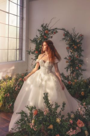 Ethereal wedding gown amid floral arch with roses and greenery in sunlit indoor garden setting.
