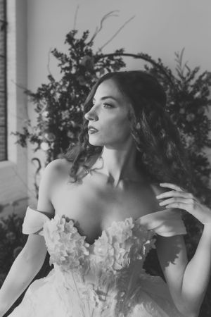 Black and white photograph of a bride in an off-shoulder wedding gown with floral details gazing to the side.