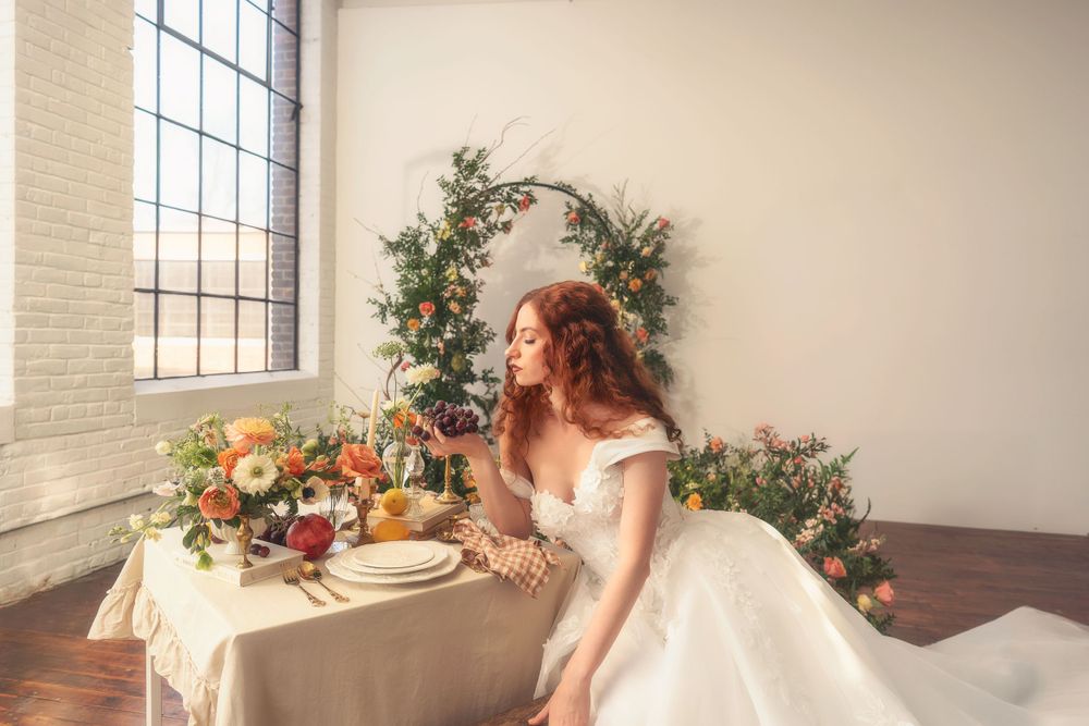 A bride in white dress sits at an elegantly decorated table with floral arrangements and arched greenery backdrop by window.