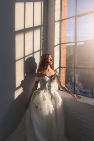 A bride in a white embellished ballgown stands by a sunlit window, casting dramatic shadows in an elegant indoor setting.