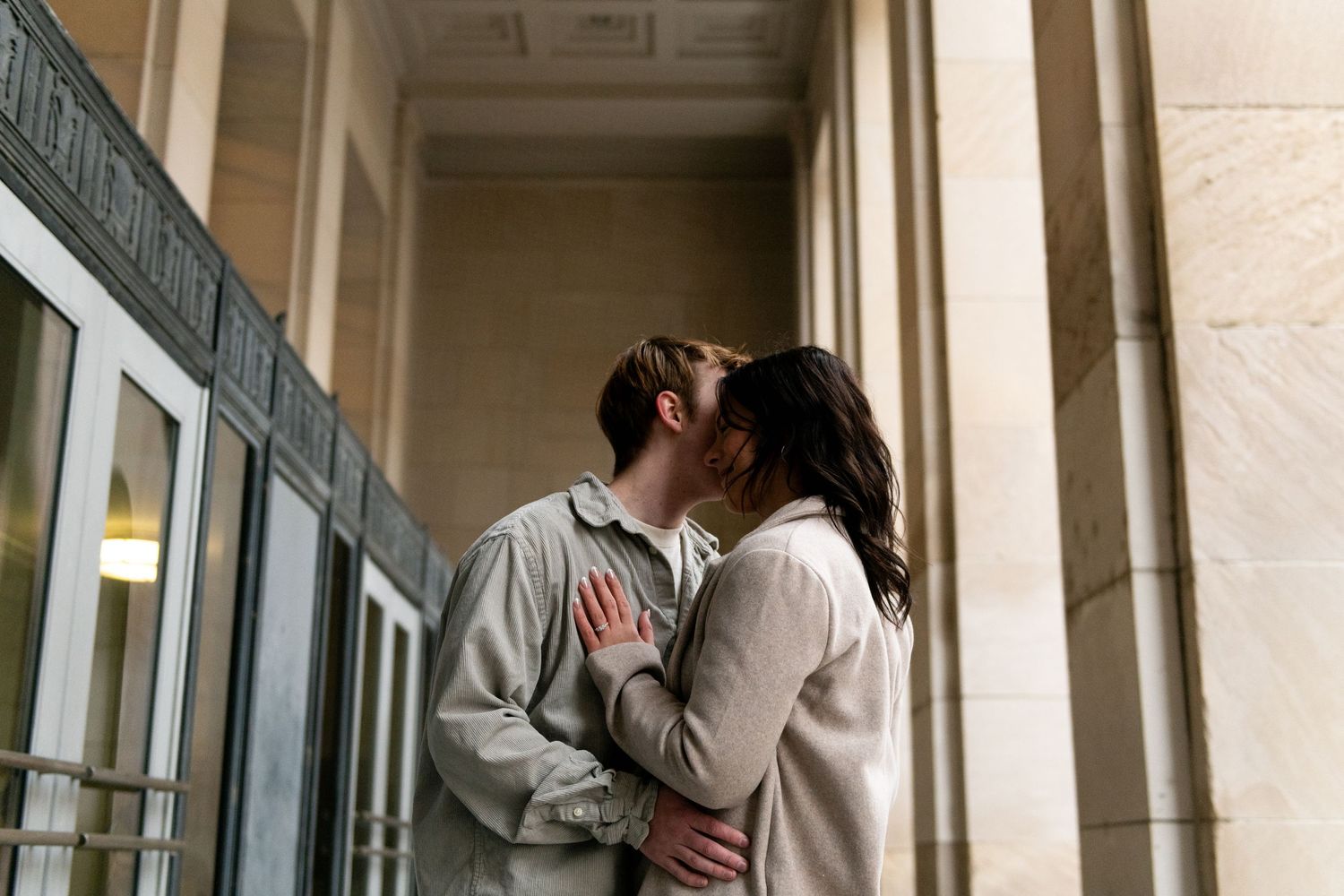 A couple shares a romantic kiss near ornate columns in an elegant indoor setting with natural lighting.