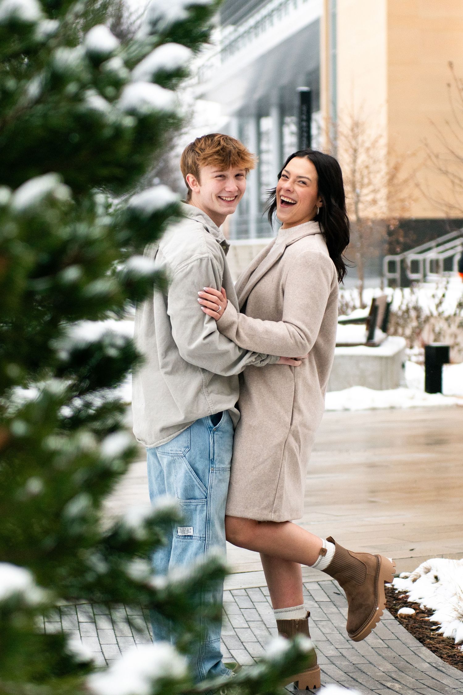 A couple embraces and laughs together on a snowy winter day, wearing cozy sweaters and casual attire.