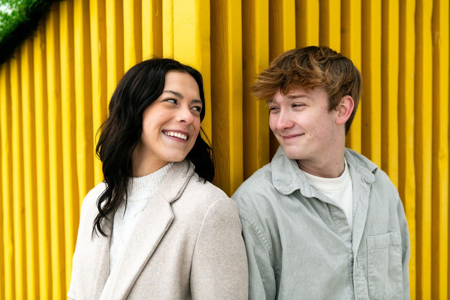 Two people laugh and smile together while standing against a bright yellow corrugated wall backdrop.