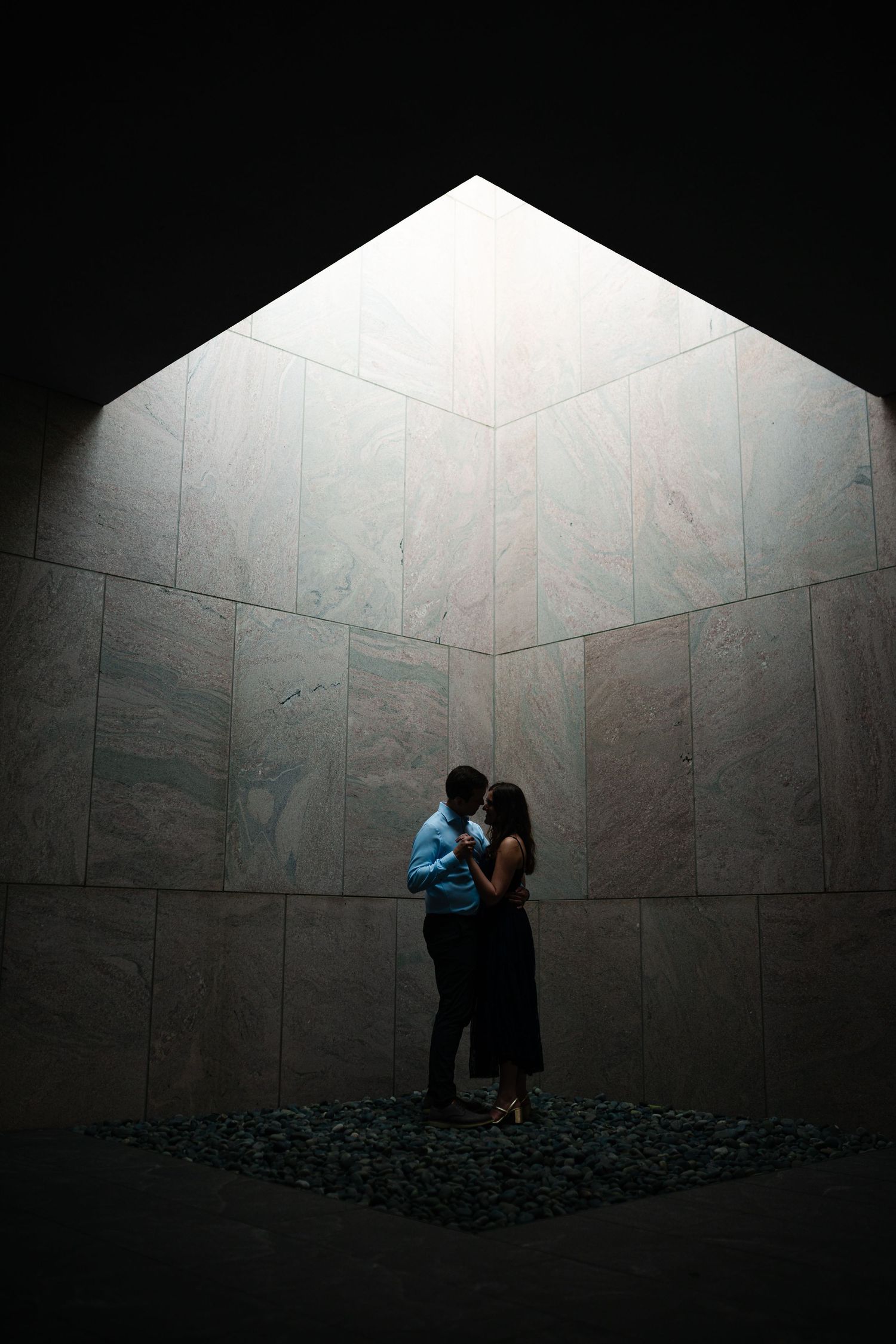 A couple stands intimately in a dramatic concrete corner illuminated by natural light from above, creating a moody atmosphere.