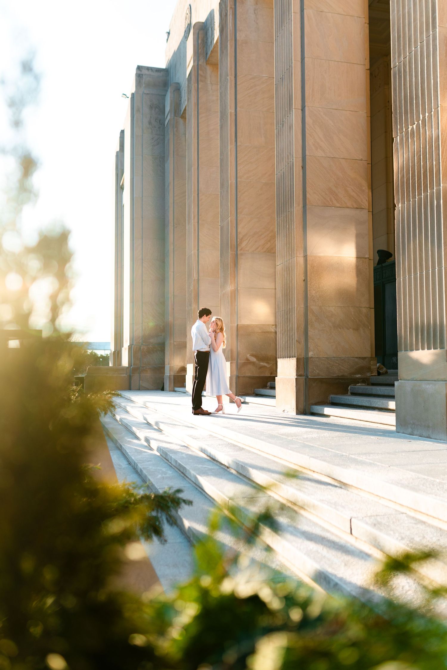 A couple shares a romantic moment between grand columns at sunset, with sunlight casting long shadows across marble steps.