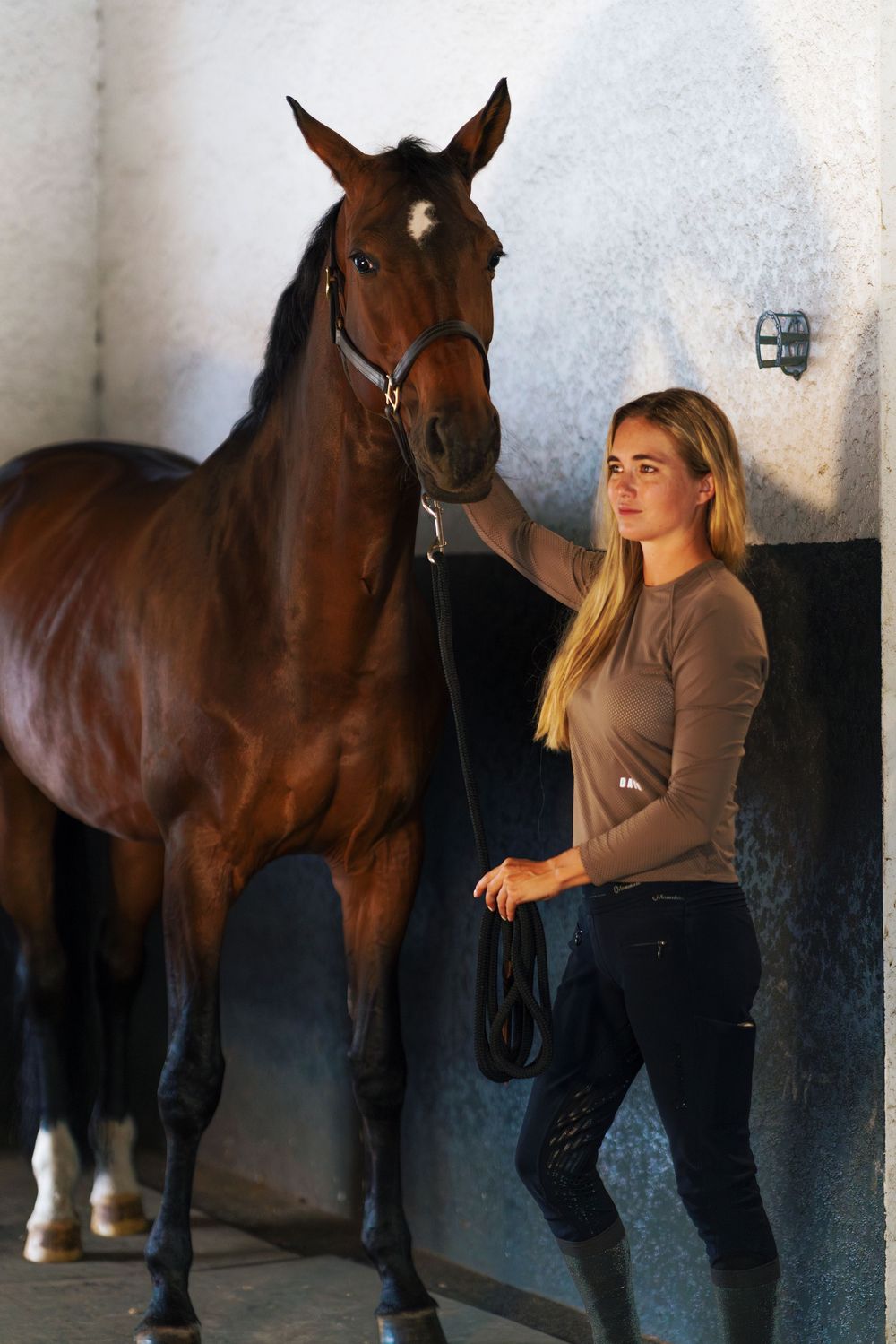 A woman in a brown sweater poses with a dark bay horse in a stable hallway against a light colored wall.