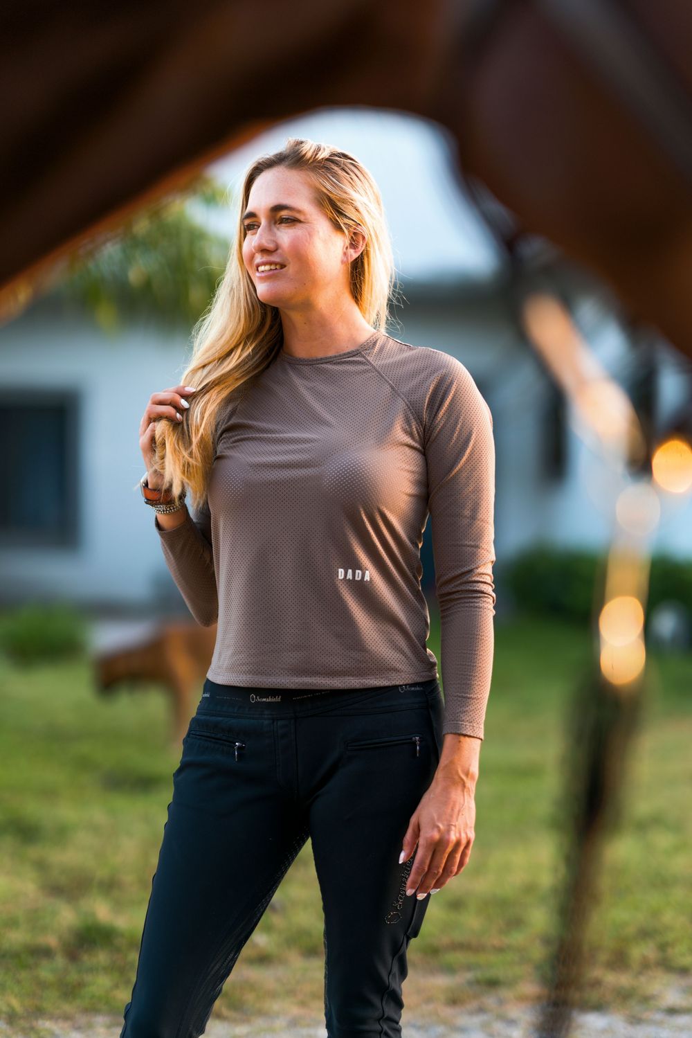 Person wearing a brown long sleeve top and dark jeans stands outdoors on grass with a house in the background.