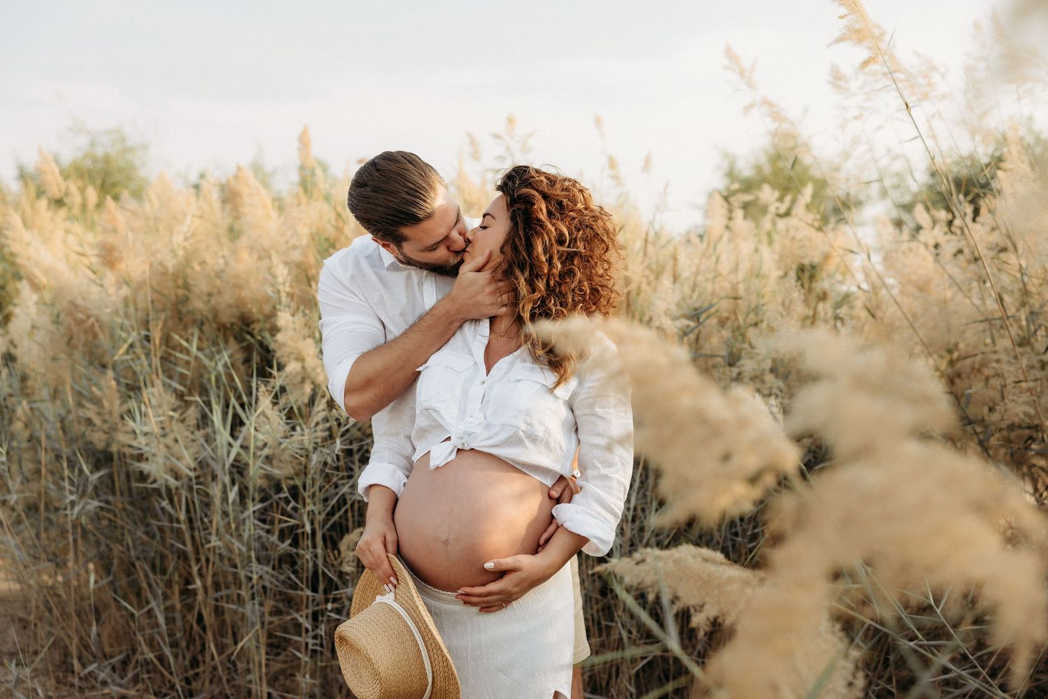 A cheerful maternity moment of a couple standing outdoors in the greenery, laughing together as the man holds the woman gently.