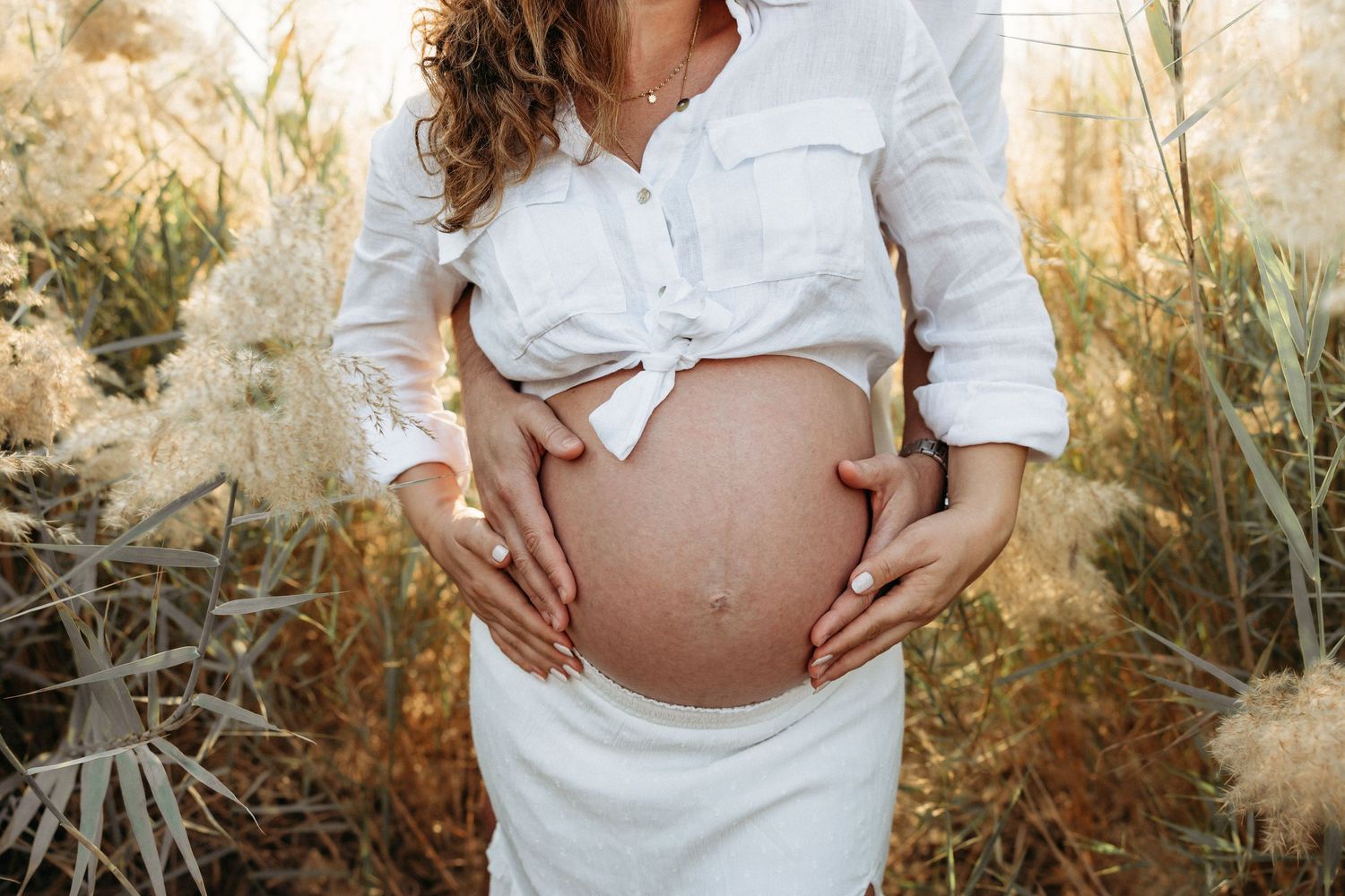 A close-up maternity portrait of a woman holding her baby bump while standing amidst golden grass, dressed in a soft white outfit.