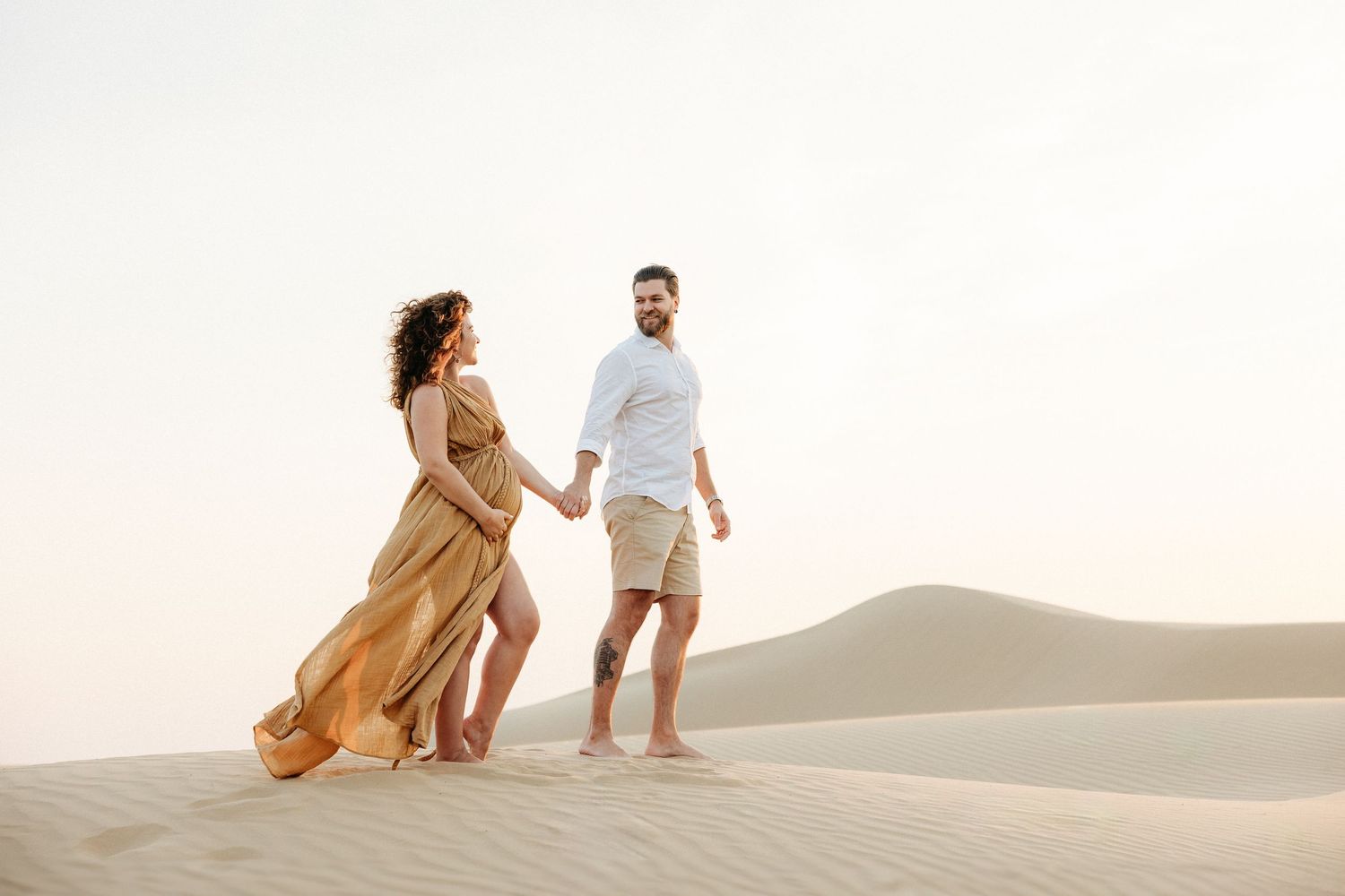 A maternity couple walking hand-in-hand on sand dunes; she wears a flowing golden dress, he a white shirt and shorts, with a serene desert.