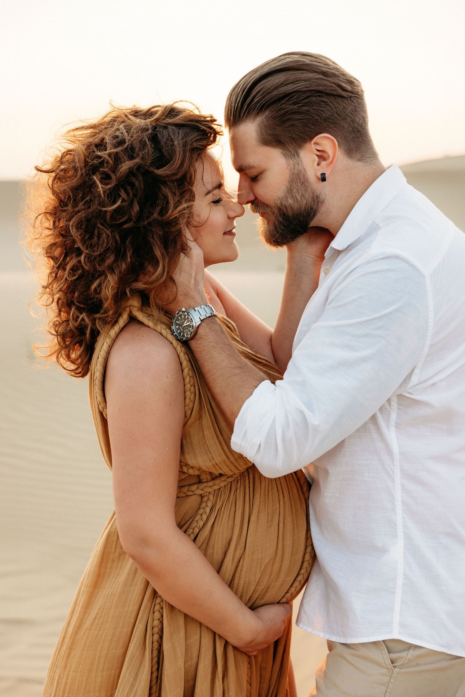 An intimate maternity portrait of a couple, with the man embracing the woman as she cradles her baby bump, set in the desert at sunset.