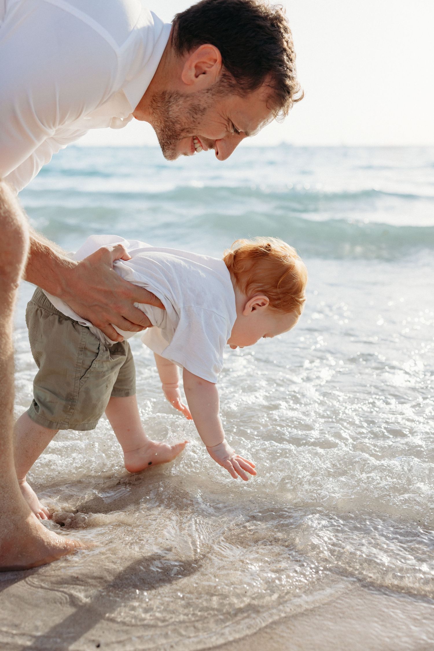 A playful father-and-son moment on the beach, with the father crouching as the toddler touches the waves for the first time.