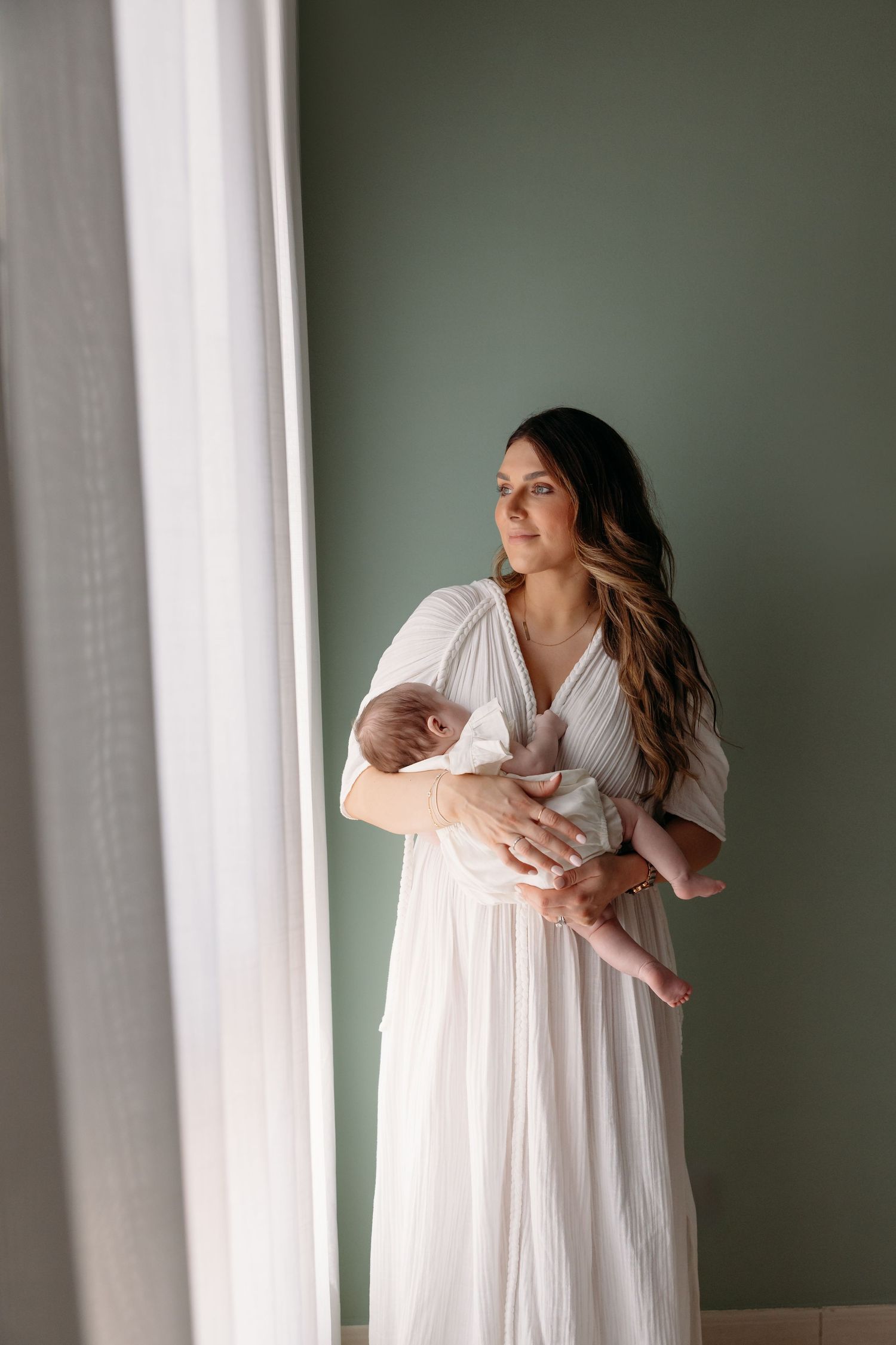 A serene indoor maternity portrait of a mother in a flowing white dress, standing near a window and cradling her newborn baby.