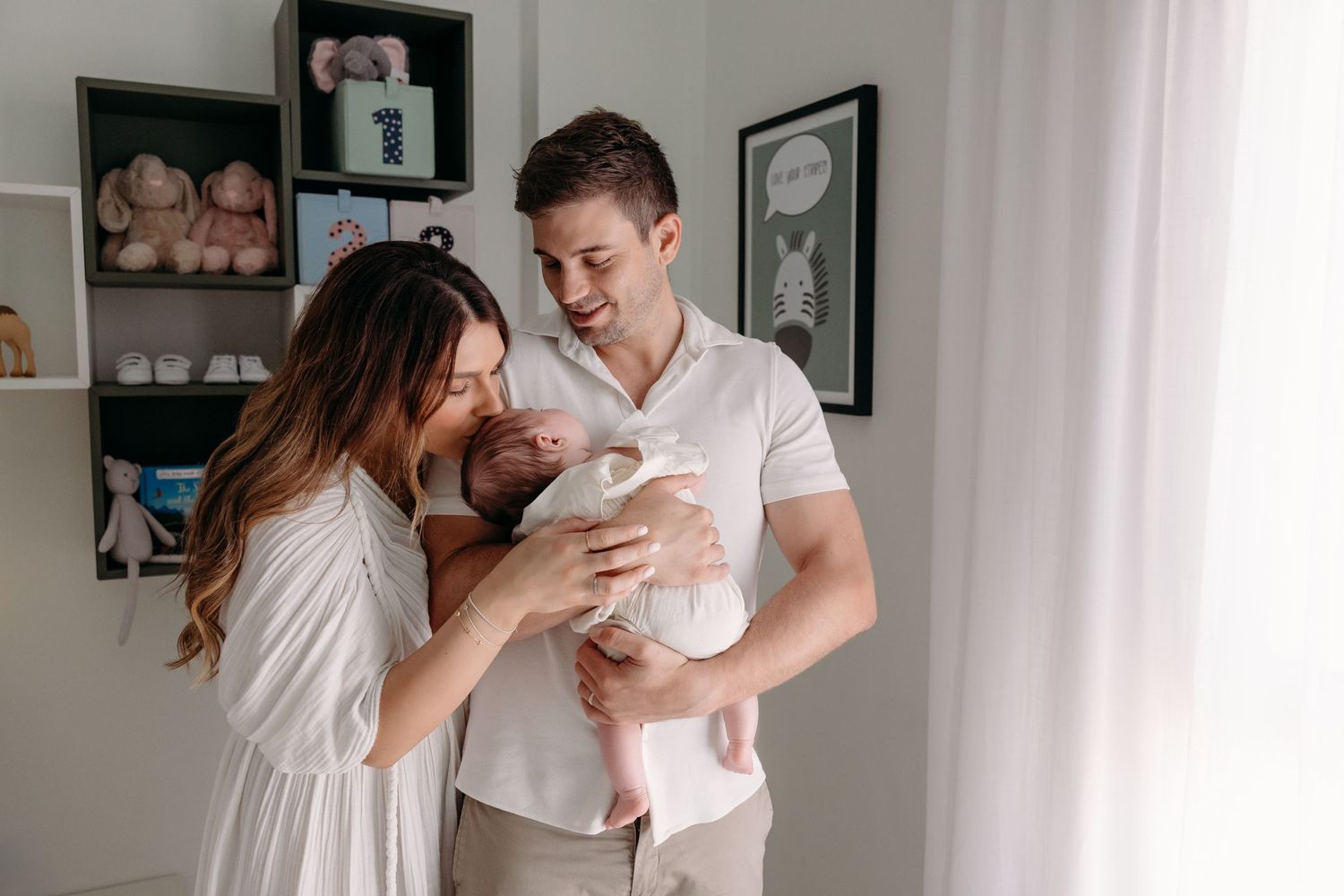 A cozy family moment in a nursery, with the parents lovingly holding their newborn baby, sharing a warm smile in a softly lit room.