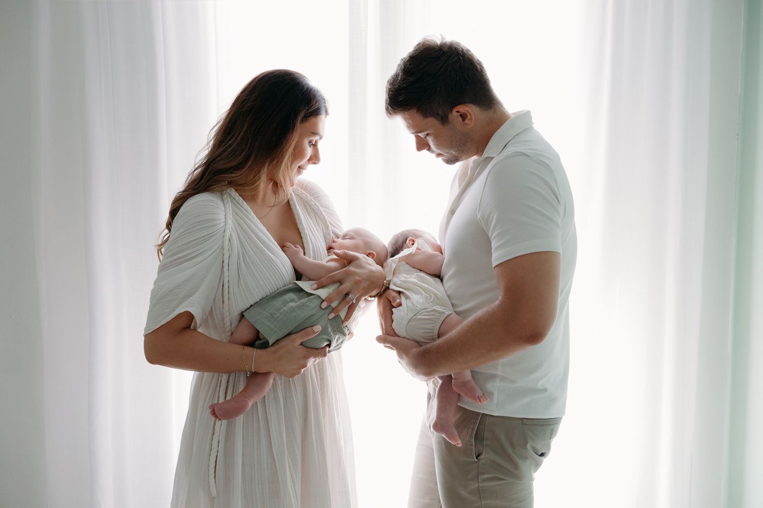An intimate indoor portrait of new parents admiring their newborn, standing close together in a softly lit nursery.