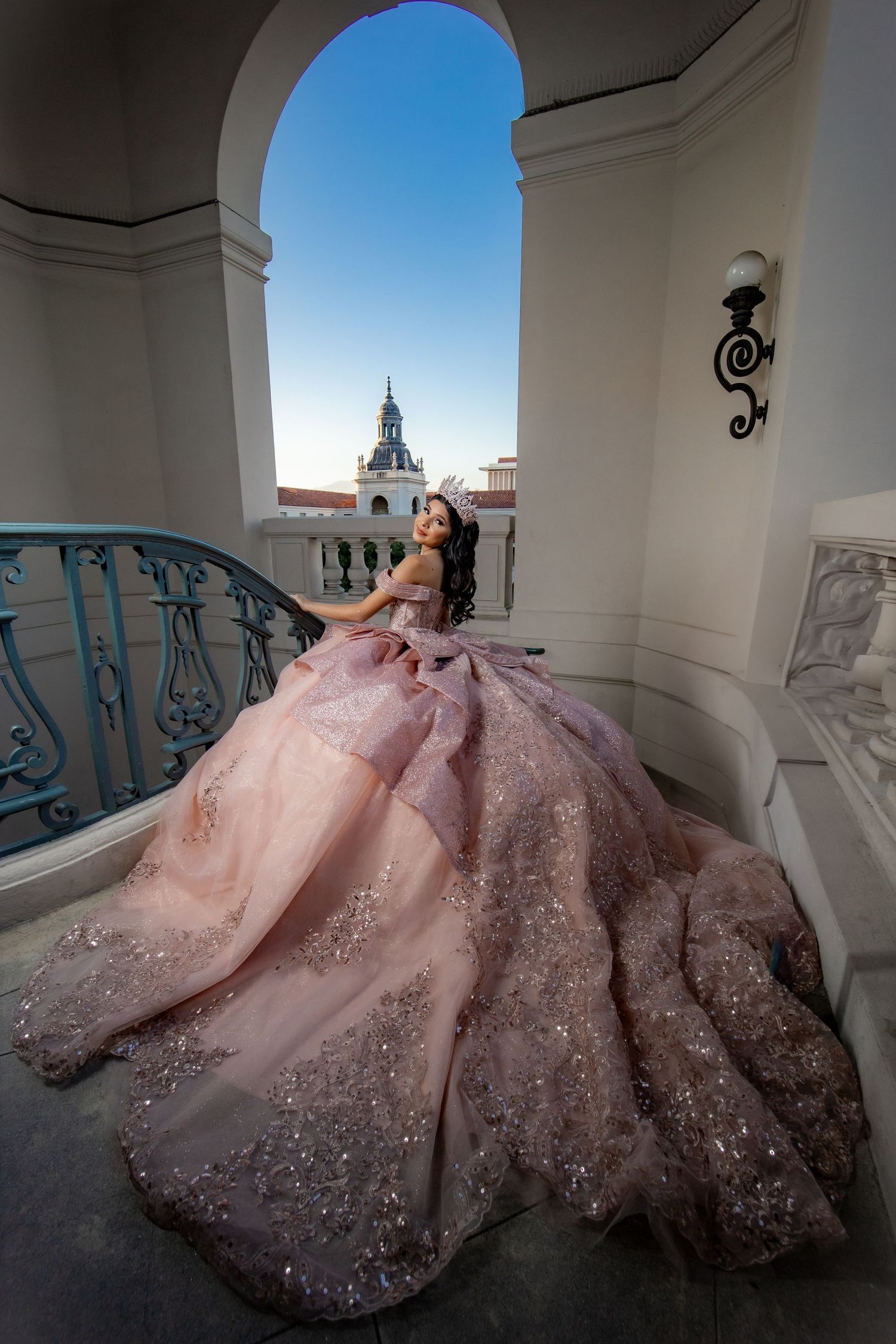 Young woman in a glittering pink quinceañera gown poses on a balcony with a city landmark in the background.