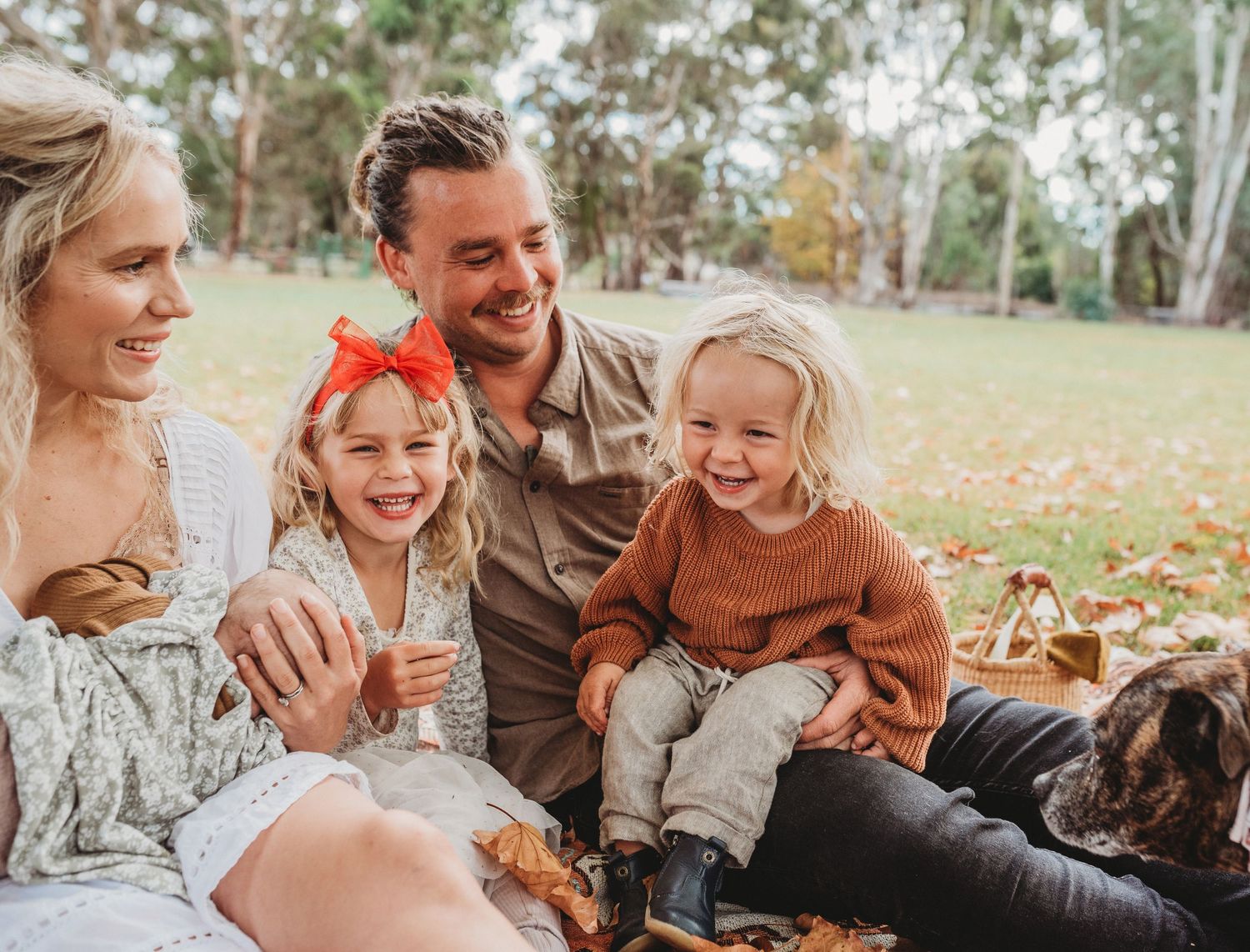 A family sits together on autumn ground surrounded by fallen leaves in a natural outdoor setting.