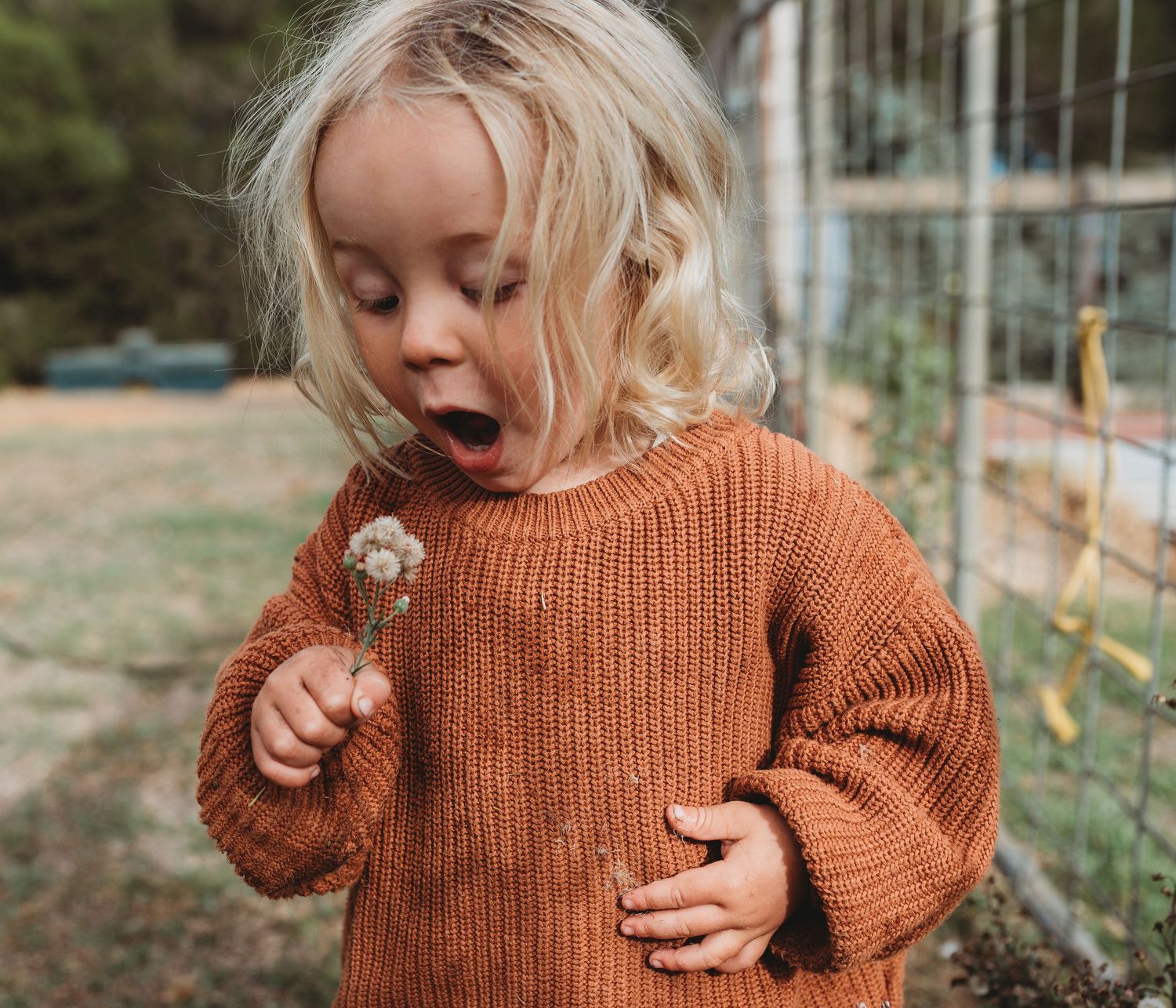 A child in an orange sweater holds a dandelion while making an excited expression outdoors.