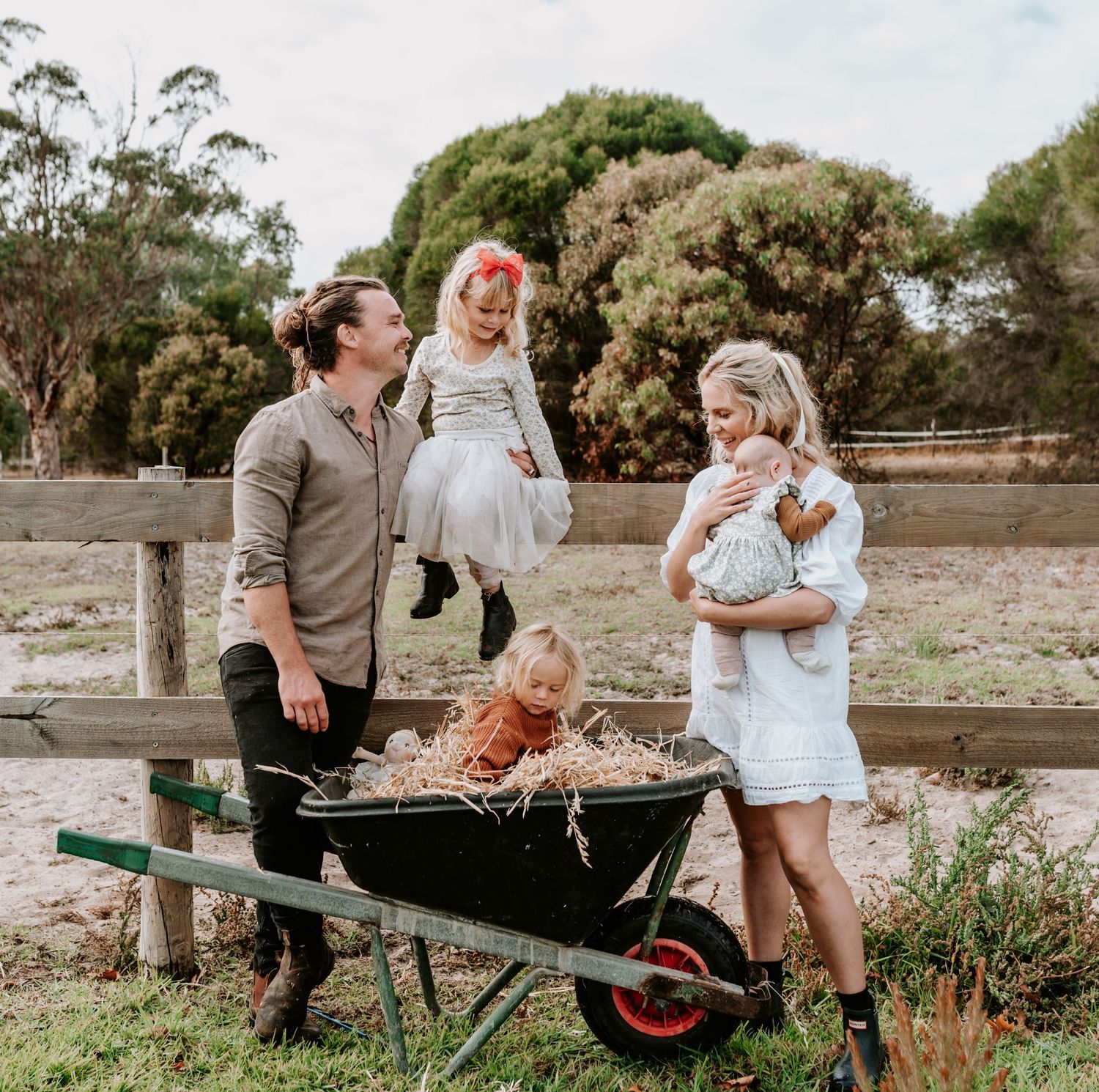 A family gathers around a wheelbarrow filled with hay in a rustic farm setting.
