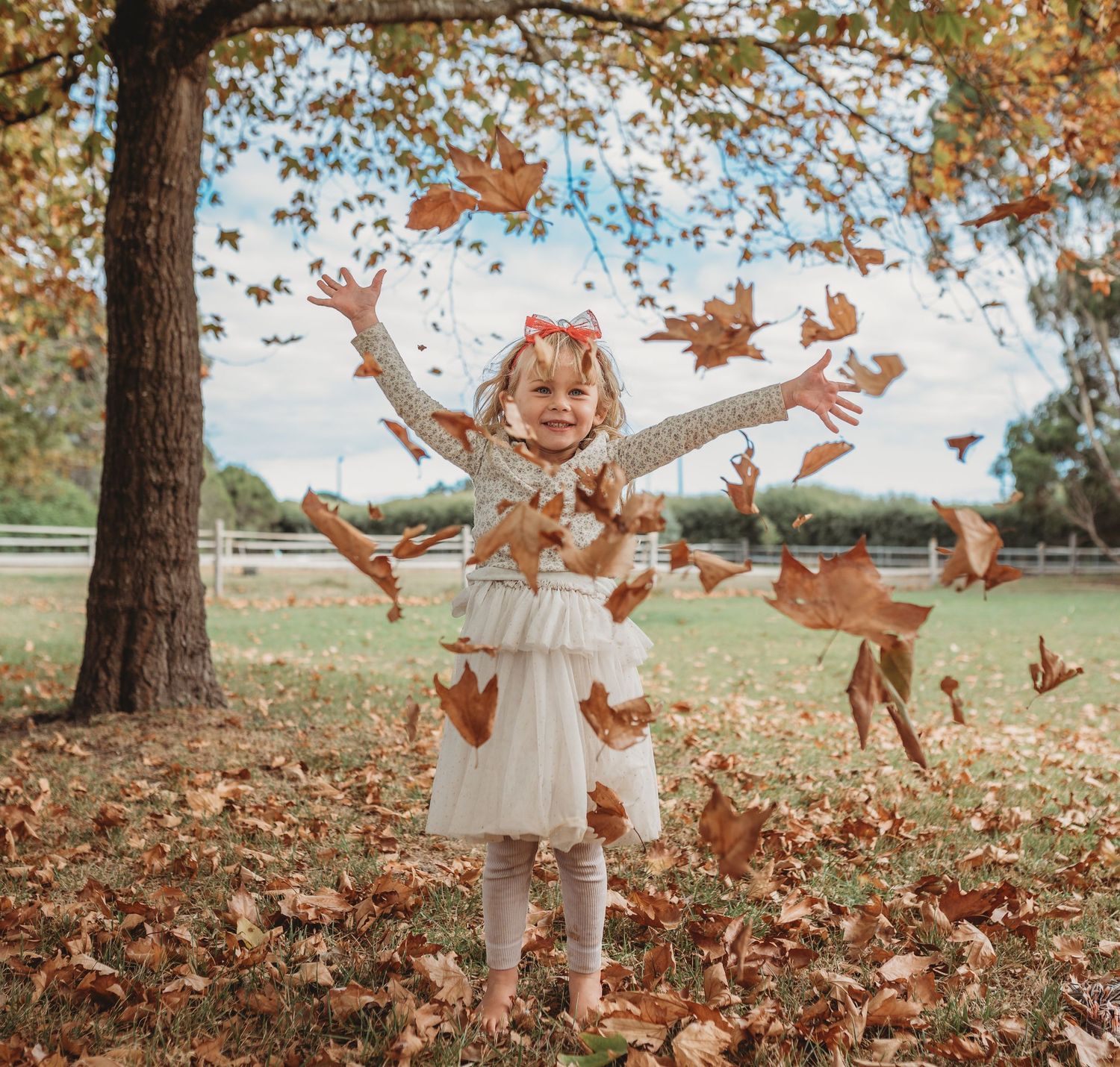 A child in a white dress throws autumn leaves in the air under a tree.