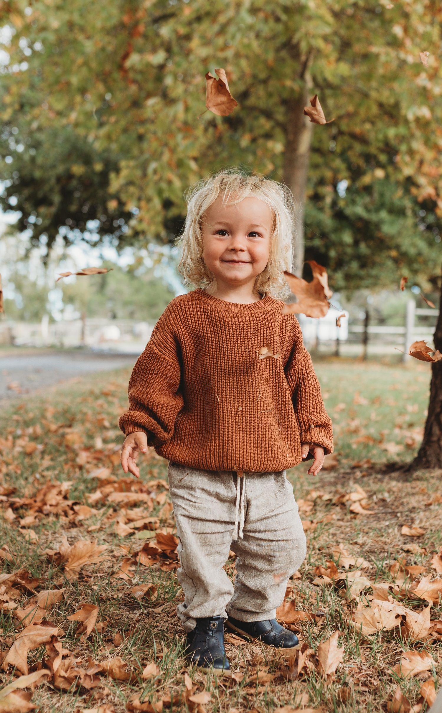 A child wearing an orange sweater stands among fallen autumn leaves.